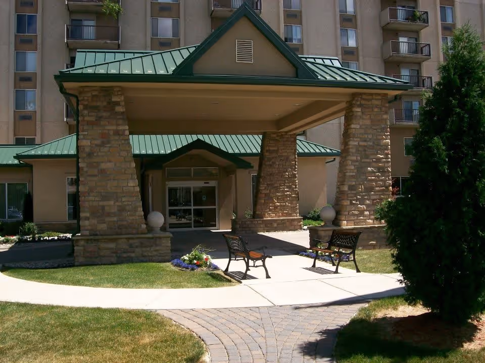 Entrance to a senior living facility with a covered drop-off area supported by stone pillars, green metal roof, two benches on a paved walkway, and a multi-story building in the background.