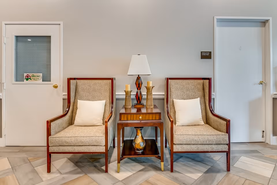 Two beige upholstered armchairs with white pillows are positioned on either side of a wooden side table with a drawer. On the table are a decorative lamp and two candle holders. The setting is indoors with a light-colored wall and two closed doors, one labeled 'Executive Director' and the other with a sign that reads 'The Country Store Oaks Only Please'. The floor has a patterned tile design.