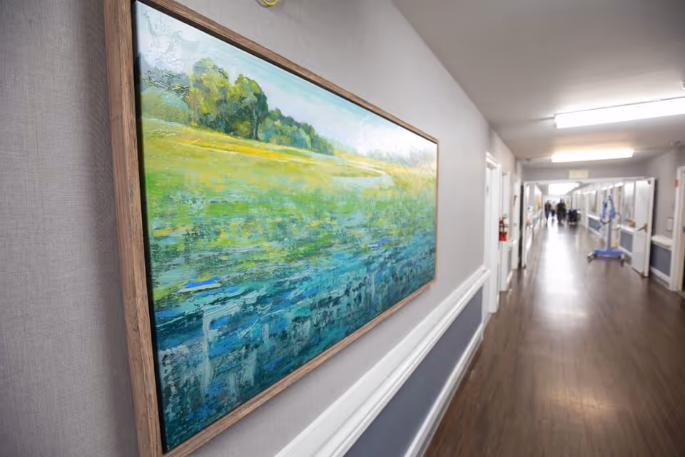 A long, well-lit hallway in a facility with wooden flooring and light gray walls. A large framed painting of a green and blue landscape is mounted on the left wall. Several doors line the hallway, and a few people are visible in the distance.
