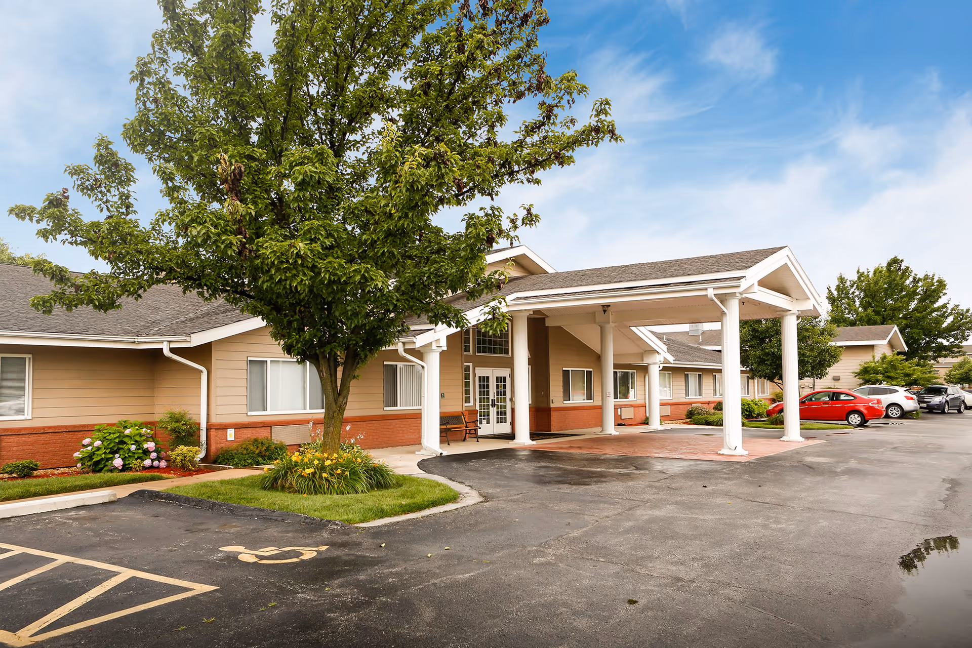 Exterior view of Gardens Independent Living facility showing a single-story building with beige and red brick walls, a covered entrance supported by white columns, a tree with green leaves, and a parking lot with several cars and a handicapped parking space.