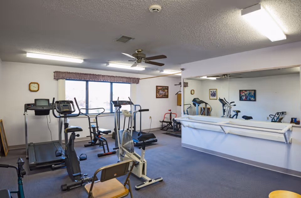 A small exercise room with various fitness equipment including a treadmill, stationary bikes, and elliptical machines. The room has a large mirror on one wall with a ballet barre attached, a ceiling fan, fluorescent lighting, and a window with a valance. There are framed pictures on the walls and a clock near the window.