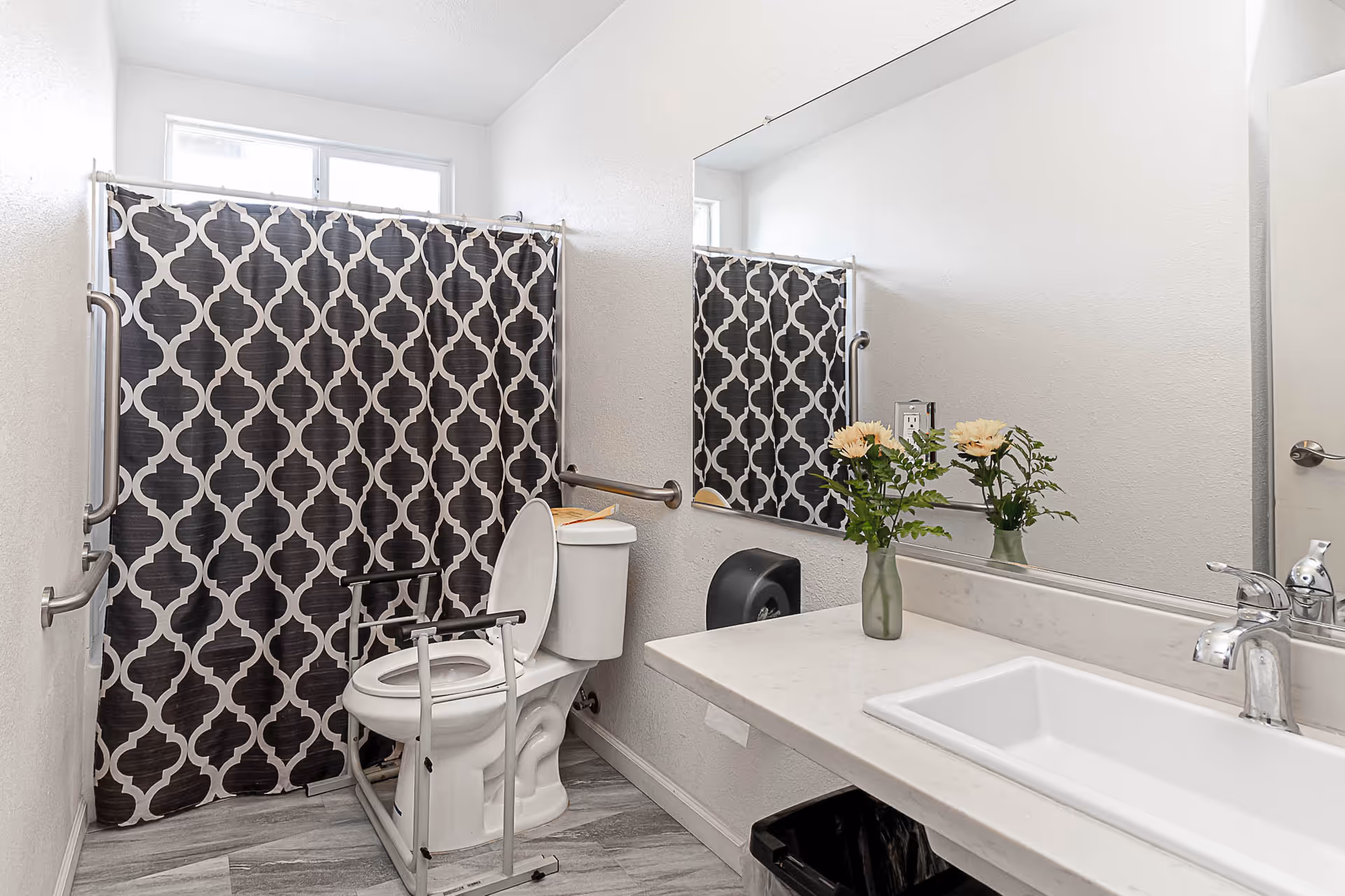 A clean bathroom with a toilet equipped with safety rails, a shower with a black and white patterned curtain, a large mirror above a white sink with a silver faucet, and a small vase with flowers on the countertop.