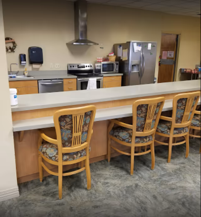A kitchen area with a long counter and four wooden chairs with floral patterned cushions. The kitchen features a stainless steel stove with a range hood, a microwave, a dishwasher, and a large refrigerator. The walls are beige, and the floor has a marbled pattern. There are paper towel dispensers and a hand sanitizer mounted on the wall.