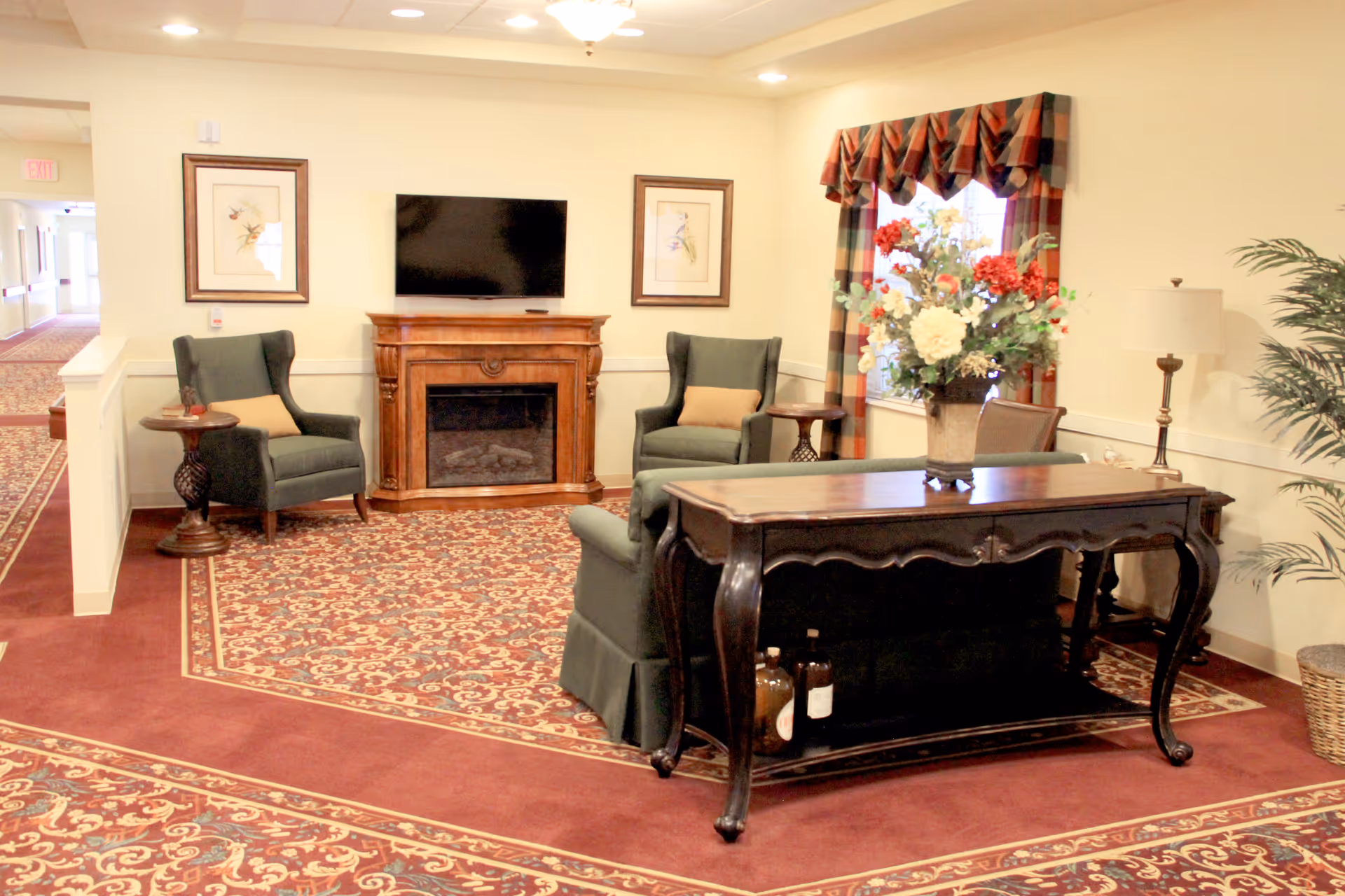 A cozy sitting area in a senior living facility with two green armchairs, a wooden electric fireplace with a TV mounted above it, a wooden console table with a floral arrangement, a floor lamp, and a window with plaid valance curtains. The floor is covered with a patterned carpet and the walls are painted light beige.