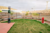 Small fenced outdoor area with green grass, a red fire hydrant, a trash can, and a signpost. In the background, there are buildings and hills under a cloudy sky.