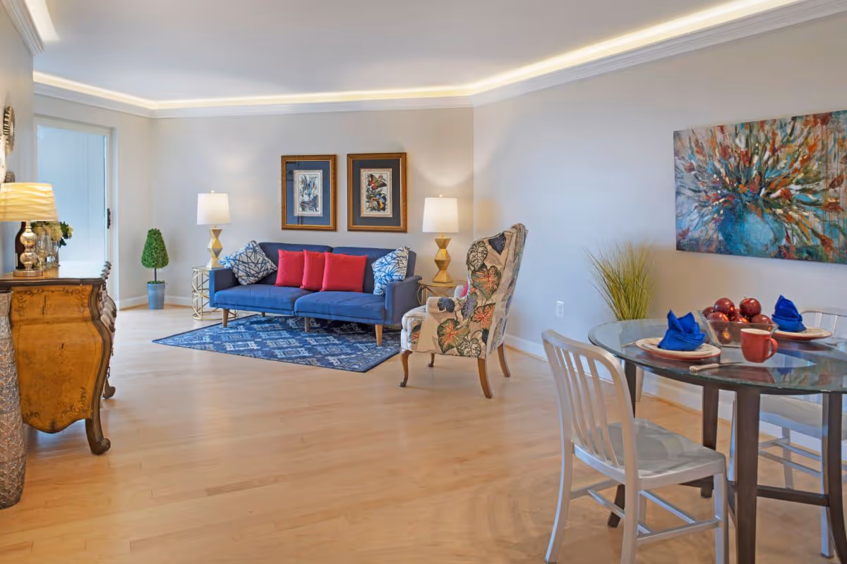 Bright open living area with a blue sofa, patterned armchair, decorative chest, and a small glass dining table set with place settings.