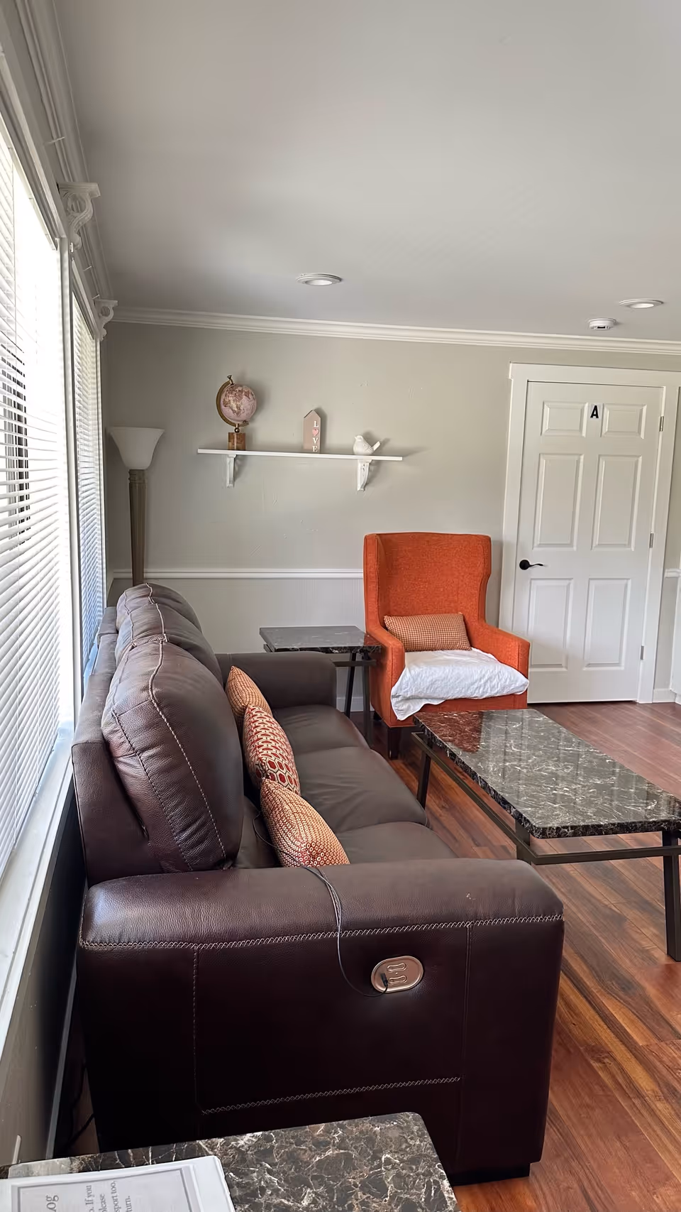 Bright living room with a brown leather sofa, patterned pillows, an orange armchair, marble-top coffee tables, and a white door labeled 'A'.