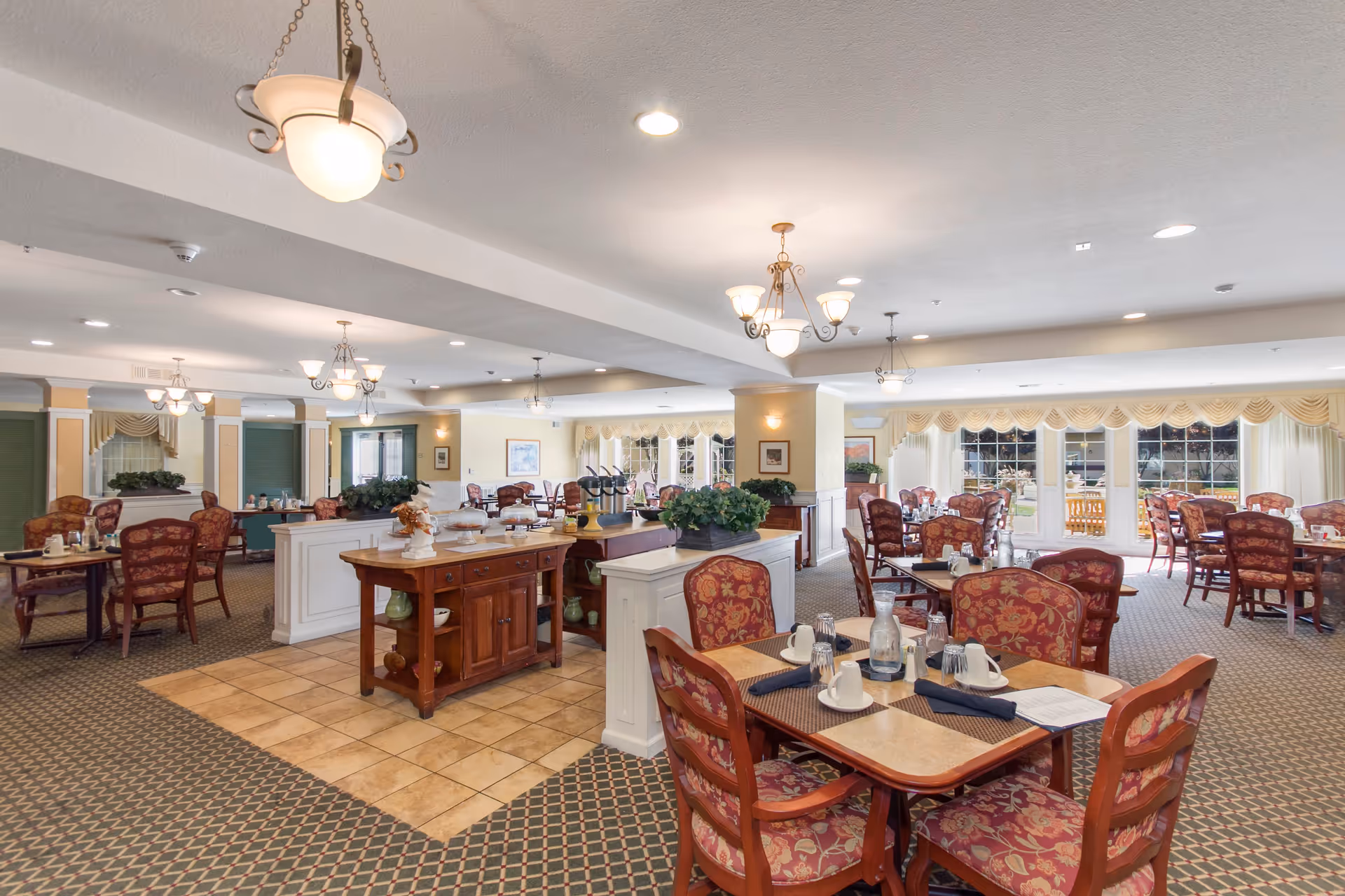 A spacious dining room in a senior living facility with multiple tables and chairs arranged neatly. The chairs have red floral upholstery, and the tables are set with cups, glasses, and napkins. The room features carpeted flooring with a tiled area in the center, decorative light fixtures hanging from the ceiling, large windows with curtains allowing natural light, and some greenery on the tables and dividers.