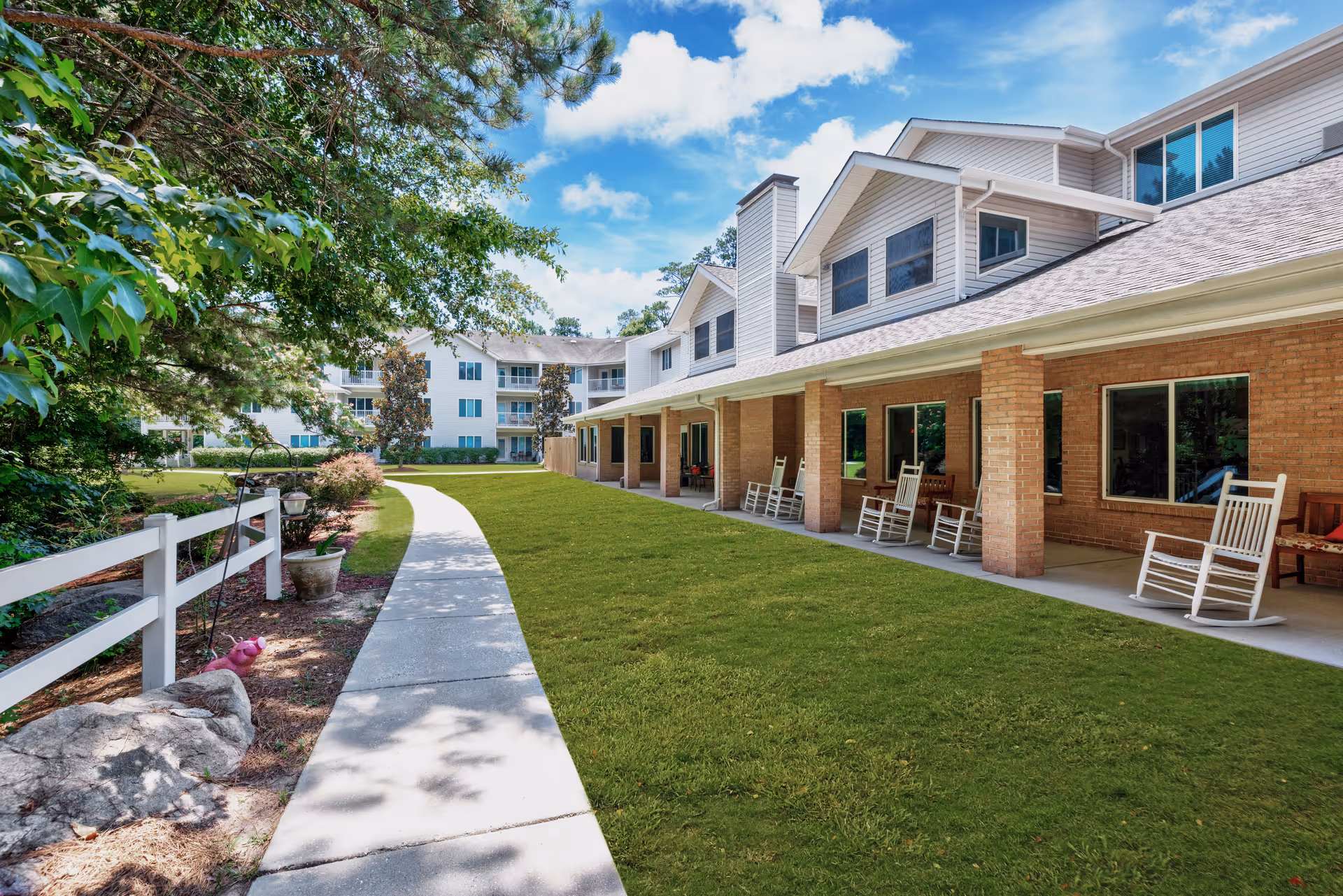 Exterior view of a senior living facility with a covered porch of rocking chairs, a walkway, and landscaped lawn.