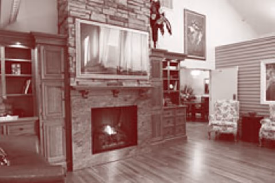A cozy living room area featuring a stone fireplace with a fire burning, a mounted flat-screen TV above it, wooden built-in shelves and cabinets on either side, two floral upholstered armchairs, and wooden flooring.