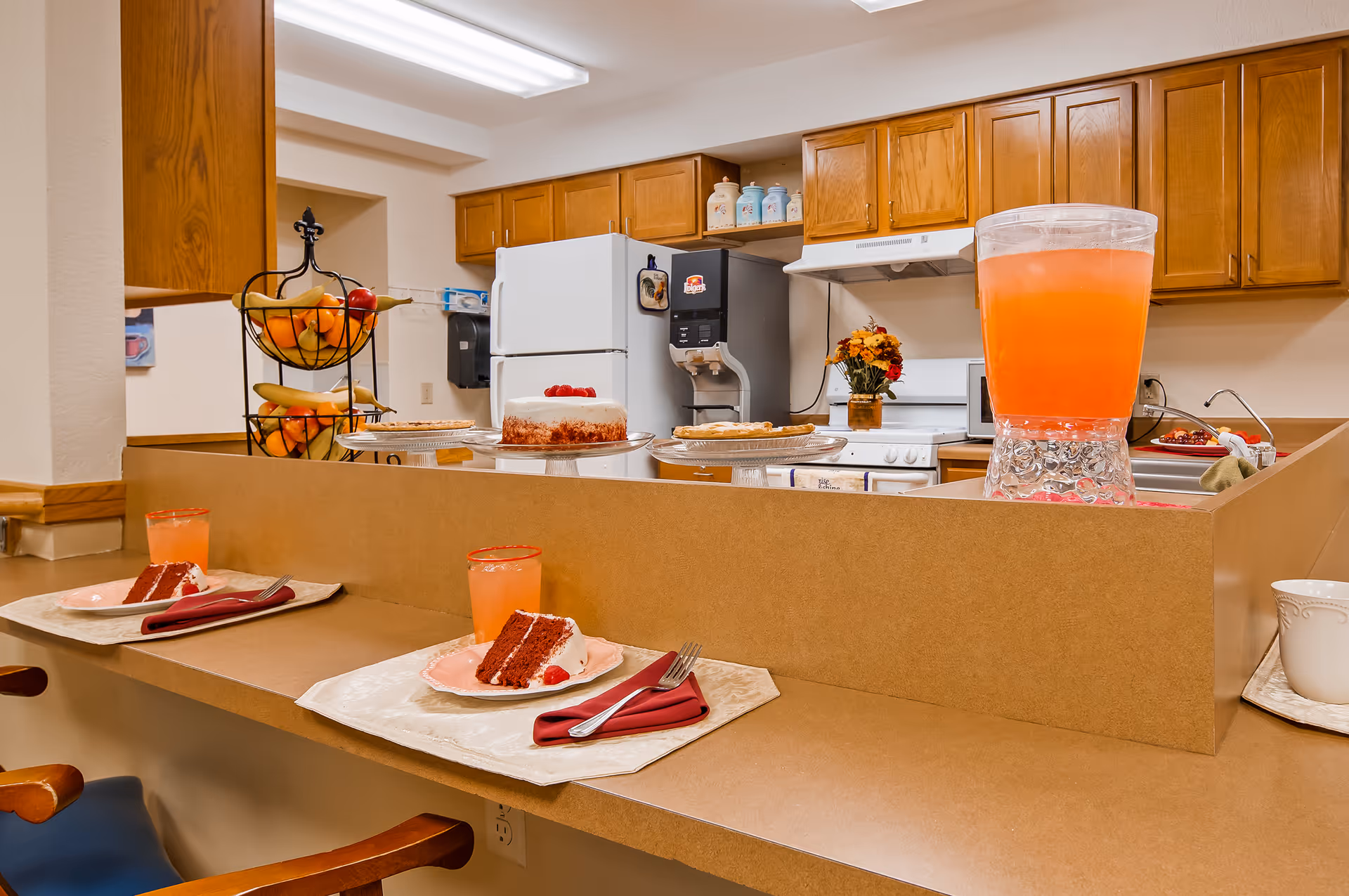 A kitchen serving counter set with slices of red velvet cake and glasses of pink lemonade, a large pitcher, fruit basket, and cabinets and appliances in the background.