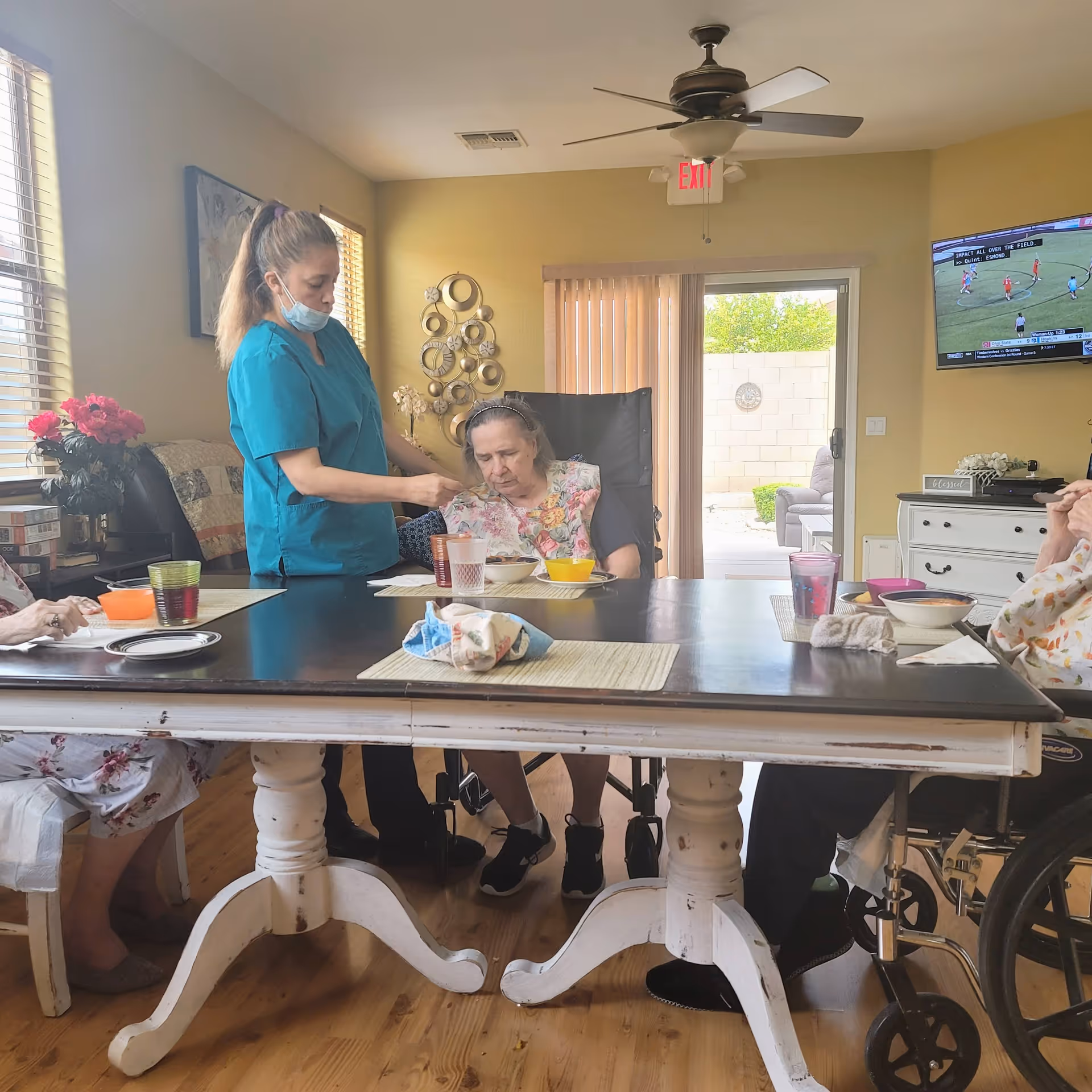 A caregiver wearing a teal uniform and face mask is assisting an elderly woman seated in a wheelchair at a dining table in a well-lit room. The table has placemats, bowls, cups, and a cloth. Other elderly individuals are partially visible seated around the table. The room has wooden flooring, a ceiling fan, a wall-mounted TV showing a sports game, and a sliding glass door leading to an outdoor area.