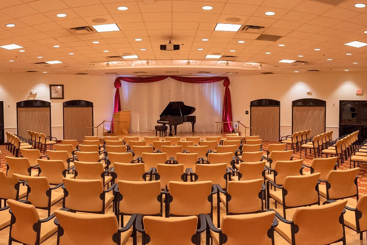 Empty assembly room with rows of beige chairs facing a stage with a grand piano and red curtains.