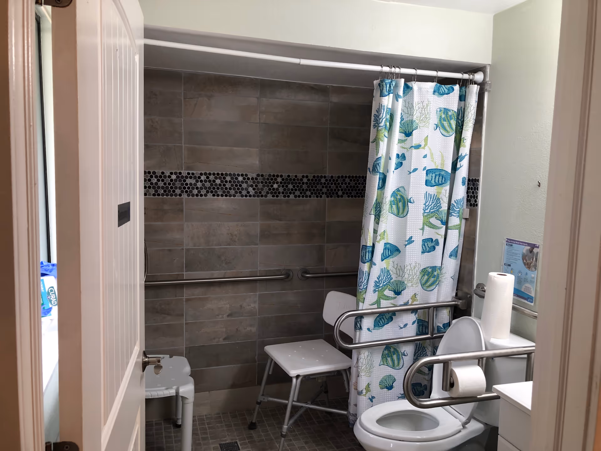 A small bathroom with a tiled walk-in shower featuring a blue and green fish-patterned shower curtain. There are two white plastic shower chairs inside the shower area. The bathroom has safety grab bars installed on the walls near the shower and toilet. A roll of paper towels is placed on top of the toilet tank, and a toilet paper roll is mounted on a grab bar next to the toilet. A hygiene instruction poster is visible on the wall above the toilet.