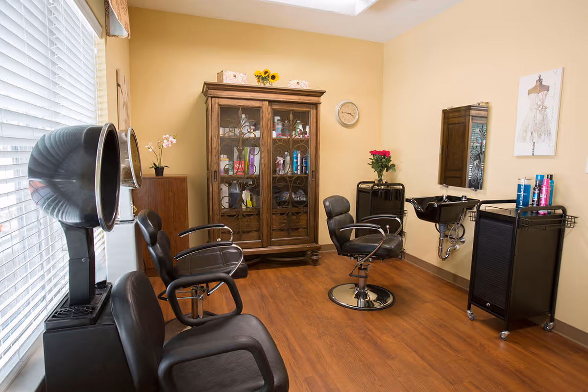 Interior of a salon room with two black salon chairs, two hair dryers, a black sink for washing hair, a wooden cabinet with glass doors containing hair products, a wall clock, and a small cart with hair care products. The room has wooden flooring and beige walls.