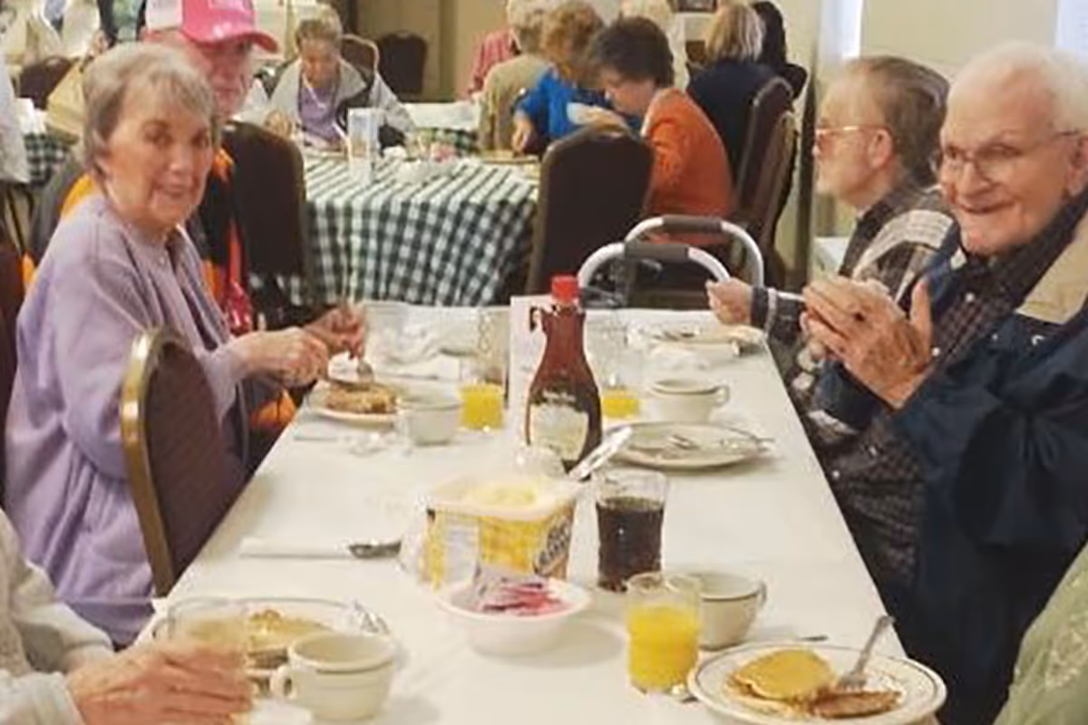 Elderly residents seated at a long table eating breakfast in a communal dining room.