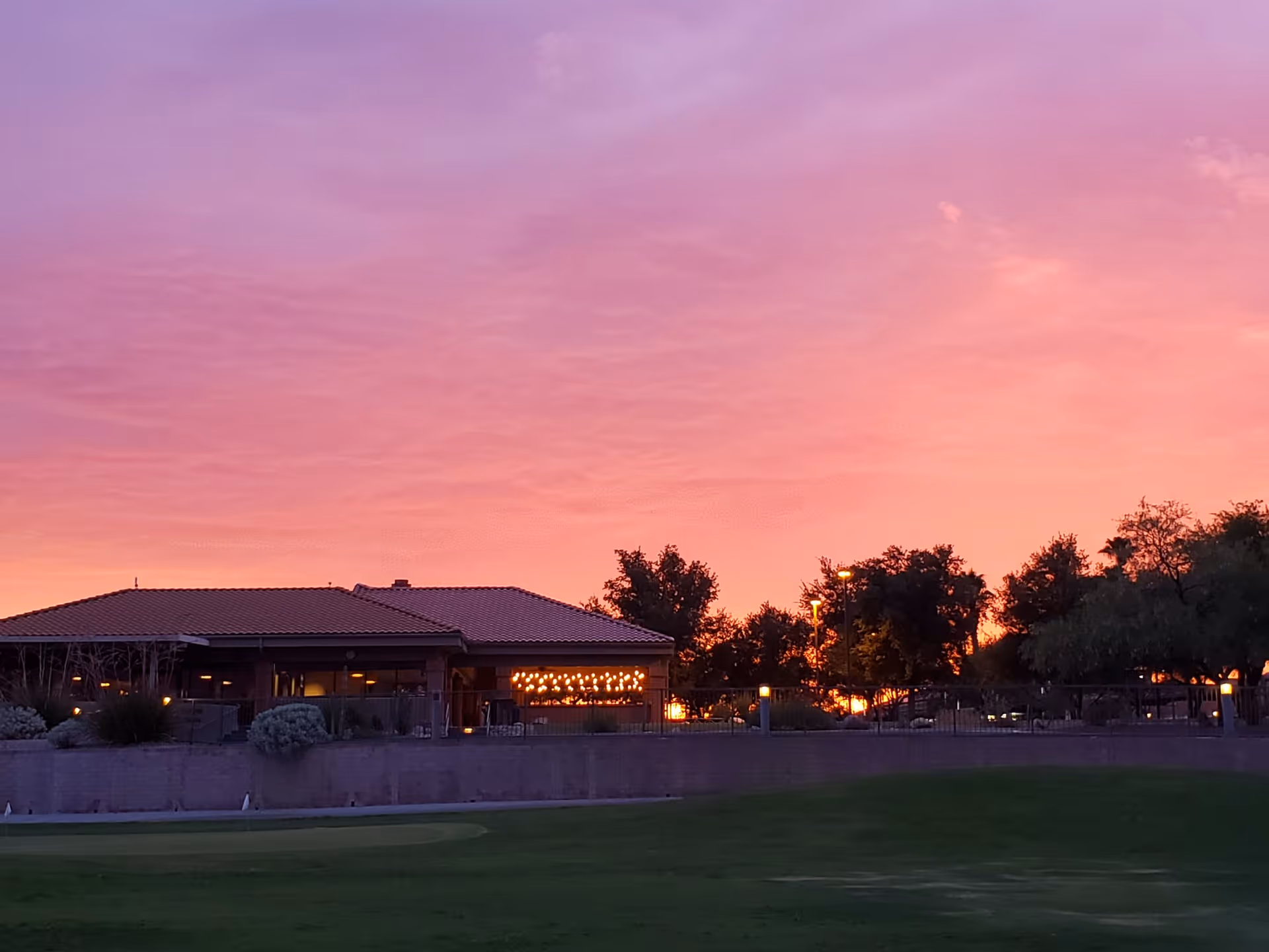 A building with a tiled roof and large windows is seen at sunset with a vibrant pink and orange sky. The building is surrounded by trees and a green lawn in the foreground.