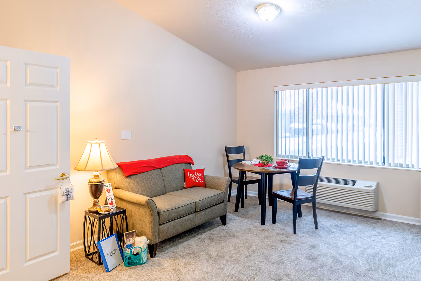 A cozy living area in an assisted living facility featuring a beige loveseat with a red throw blanket and a red pillow that says 'Love You More.' Next to the loveseat is a small black side table with a lamp, a card, and some personal items. In the background, there is a round wooden table with two chairs near a large window with vertical blinds. The room has light-colored walls and carpeted flooring.