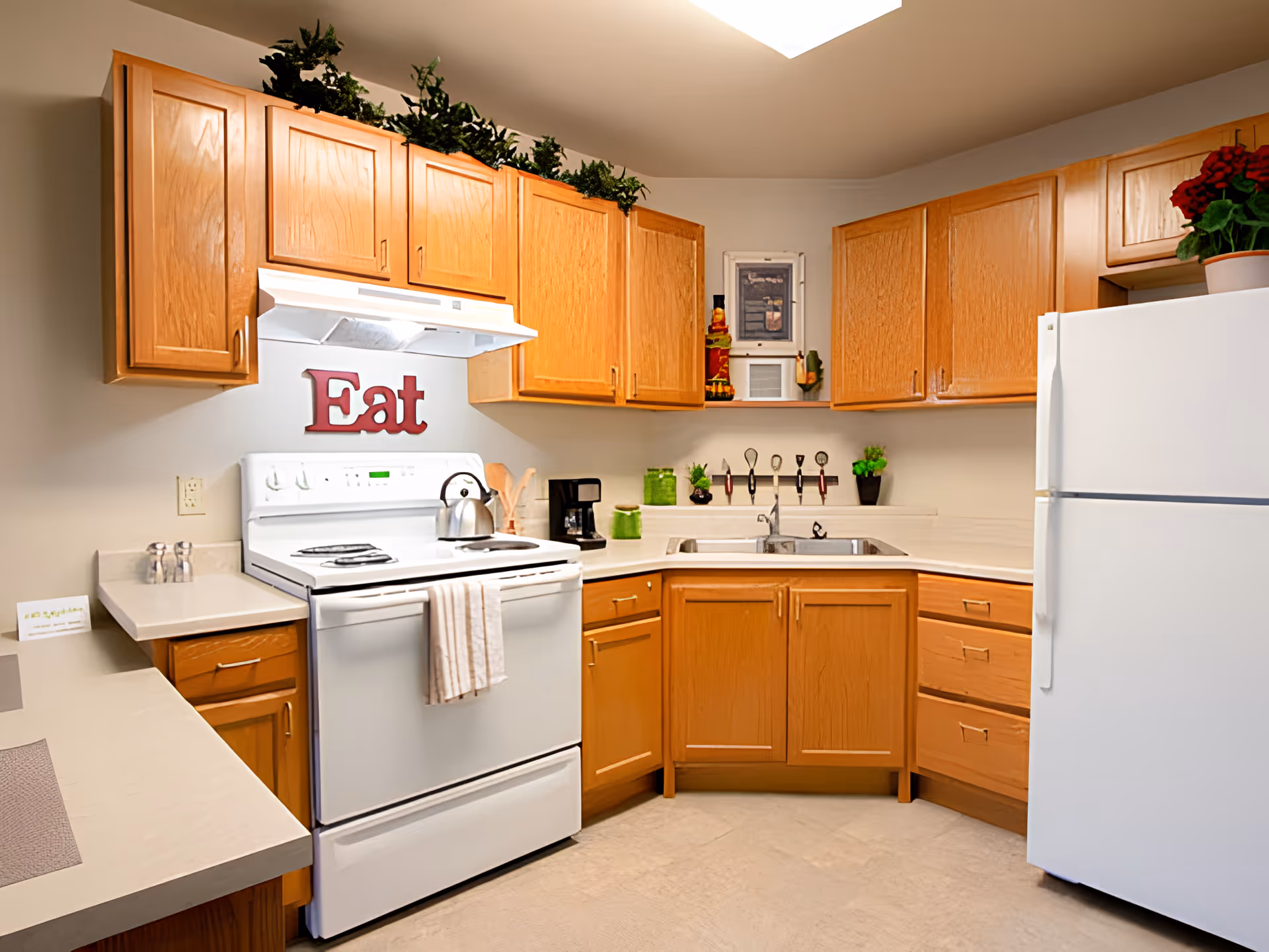 A clean kitchen with wooden cabinets, a white electric stove with a kettle on top, a white refrigerator, a double sink, and various kitchen utensils and small plants on the countertop. The word 'Eat' is displayed on the wall above the stove.