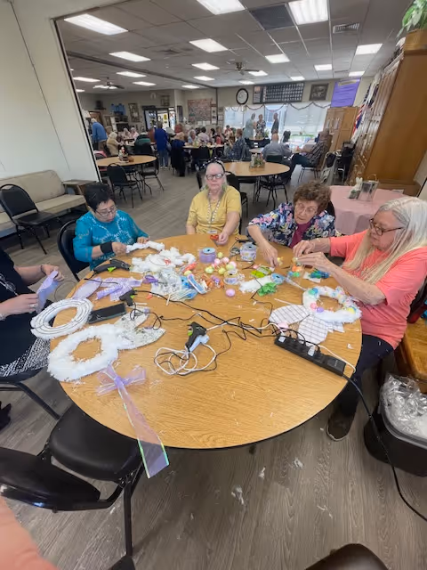 A group of elderly women sitting around a large round table engaged in a craft activity, making decorative wreaths with various materials like ribbons, faux flowers, and glue guns. The room is a communal space with other tables and people in the background, suggesting a social or recreational area in an assisted living facility.