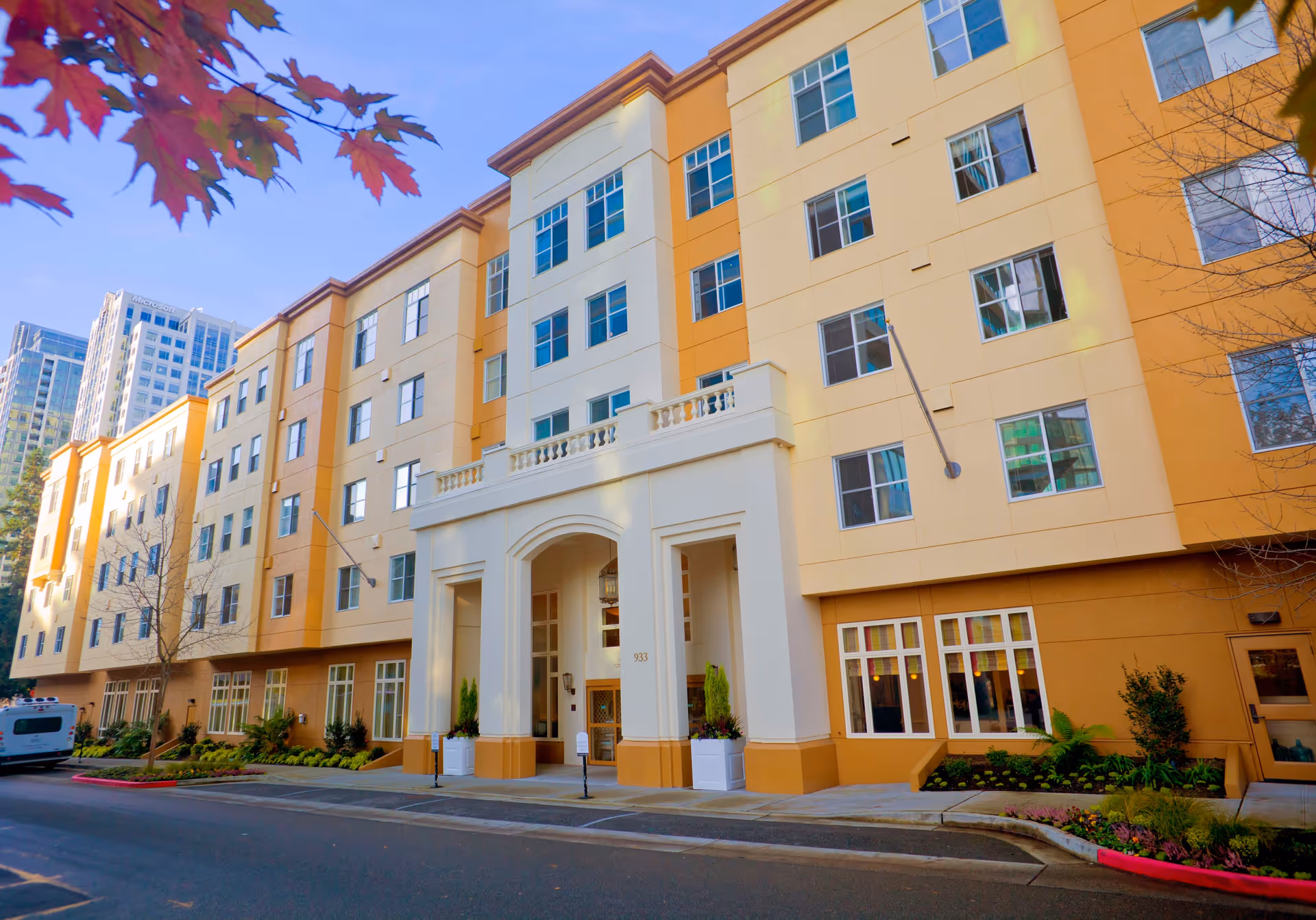 Exterior view of a multi-story senior living facility building with a beige and orange facade, large windows, and a prominent entrance with white columns and decorative railings. There are some plants and small trees along the sidewalk and a vehicle parked on the street.