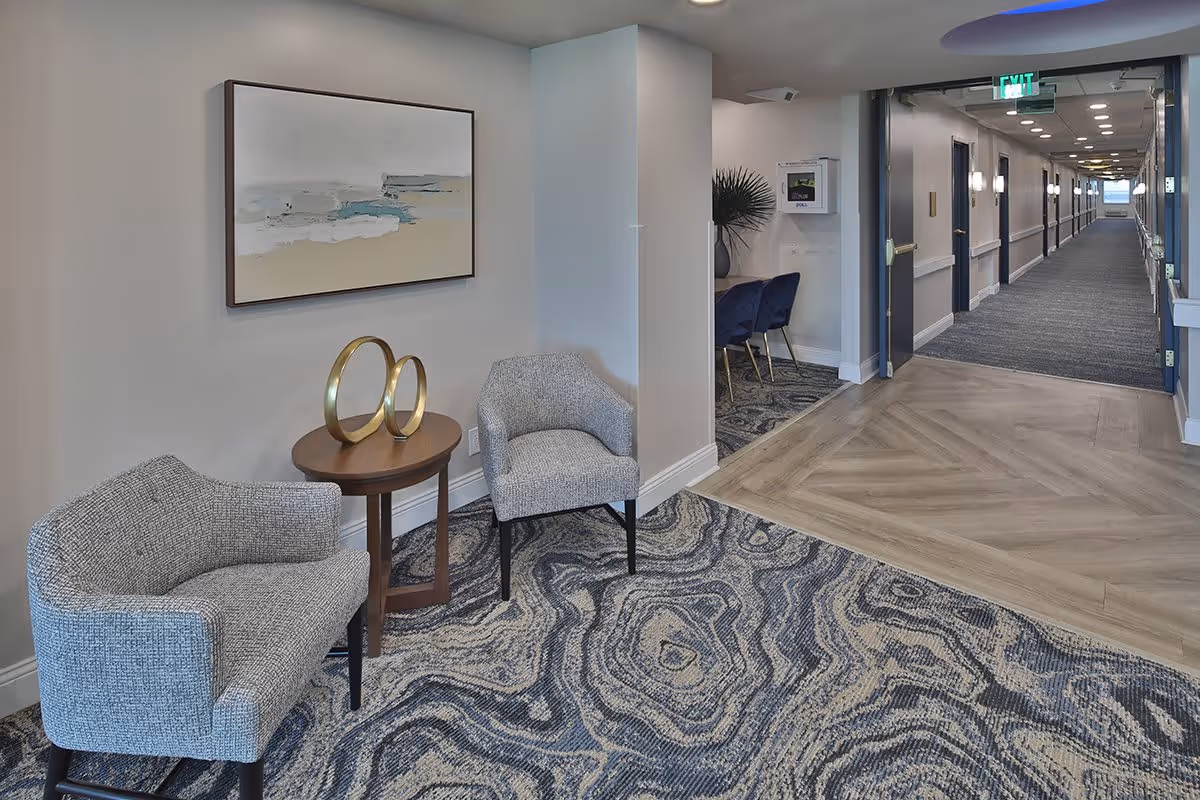A modern interior hallway area with two gray upholstered chairs and a small round wooden table between them. On the table are two decorative gold rings. Above the chairs hangs an abstract painting with neutral tones. The floor has a patterned carpet transitioning to wood flooring leading into a long corridor with multiple doors and wall-mounted lights.