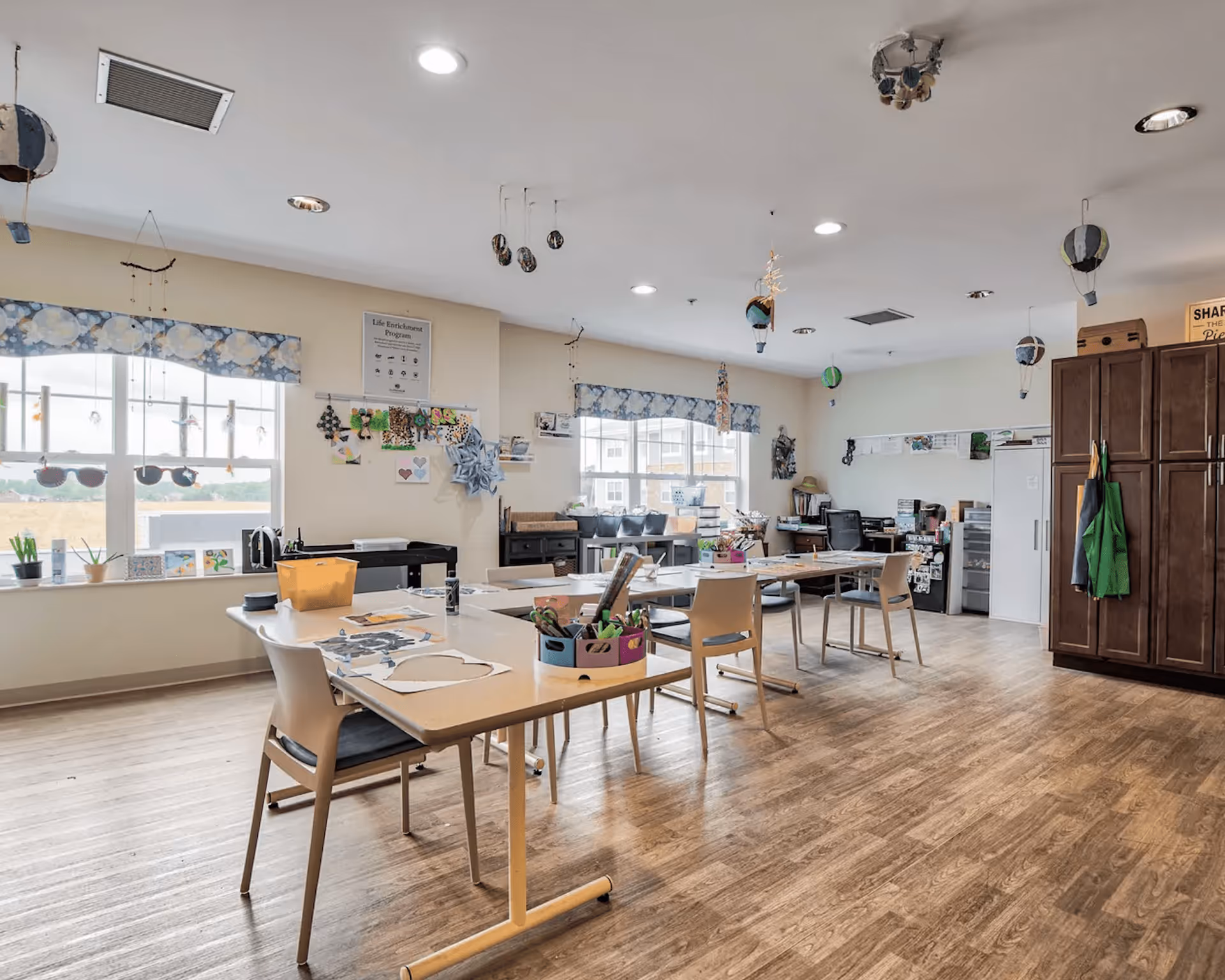 A bright and spacious activity room with wooden flooring and large windows letting in natural light. Several tables and chairs are arranged in the room, with craft supplies and decorations on the tables. The walls have various decorations, including hanging hot air balloons and artwork. There is a large cabinet with aprons hanging on the side and office equipment in the background.