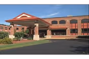 Exterior view of a brick building with a covered entrance supported by columns, surrounded by a paved driveway and some greenery under a clear sky.