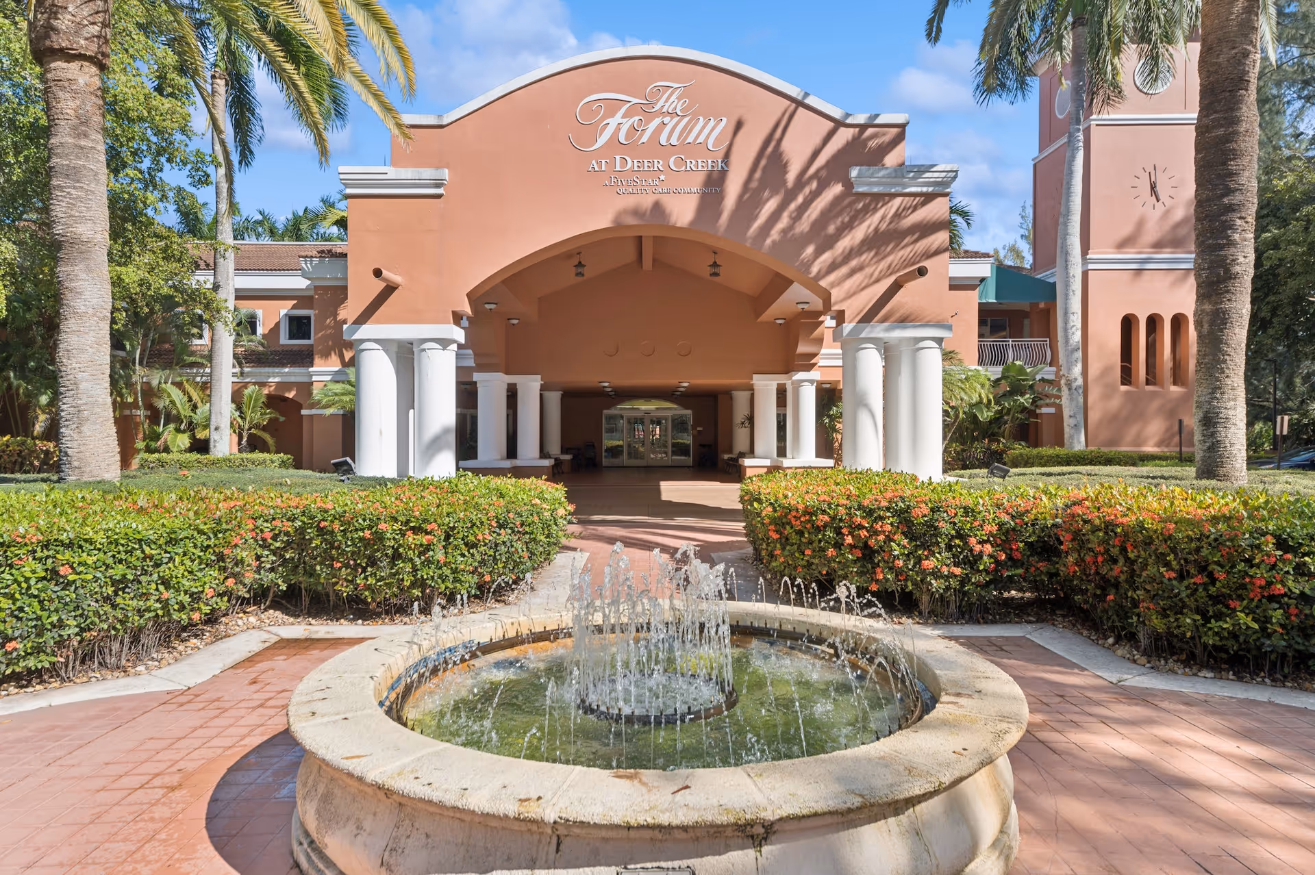 Front exterior view of The Forum at Deer Creek building with a circular water fountain in the foreground, surrounded by greenery and palm trees under a blue sky.