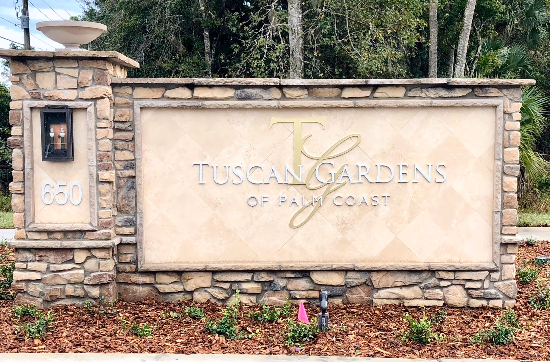 Stone and stucco entrance sign for Tuscan Gardens of Palm Coast Senior Living with the address number 650 on the left side, surrounded by landscaping and trees in the background.