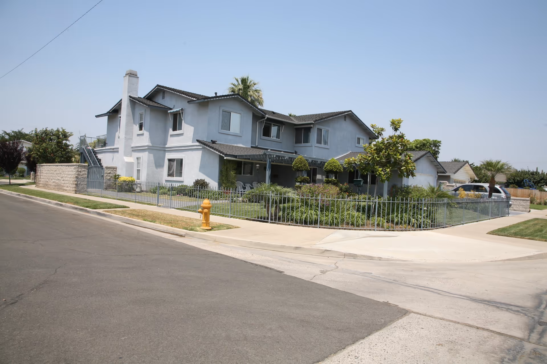 Exterior view of a two-story residential building with a light gray facade, surrounded by a metal fence and landscaped greenery, located on a corner lot with a sidewalk and a yellow fire hydrant.