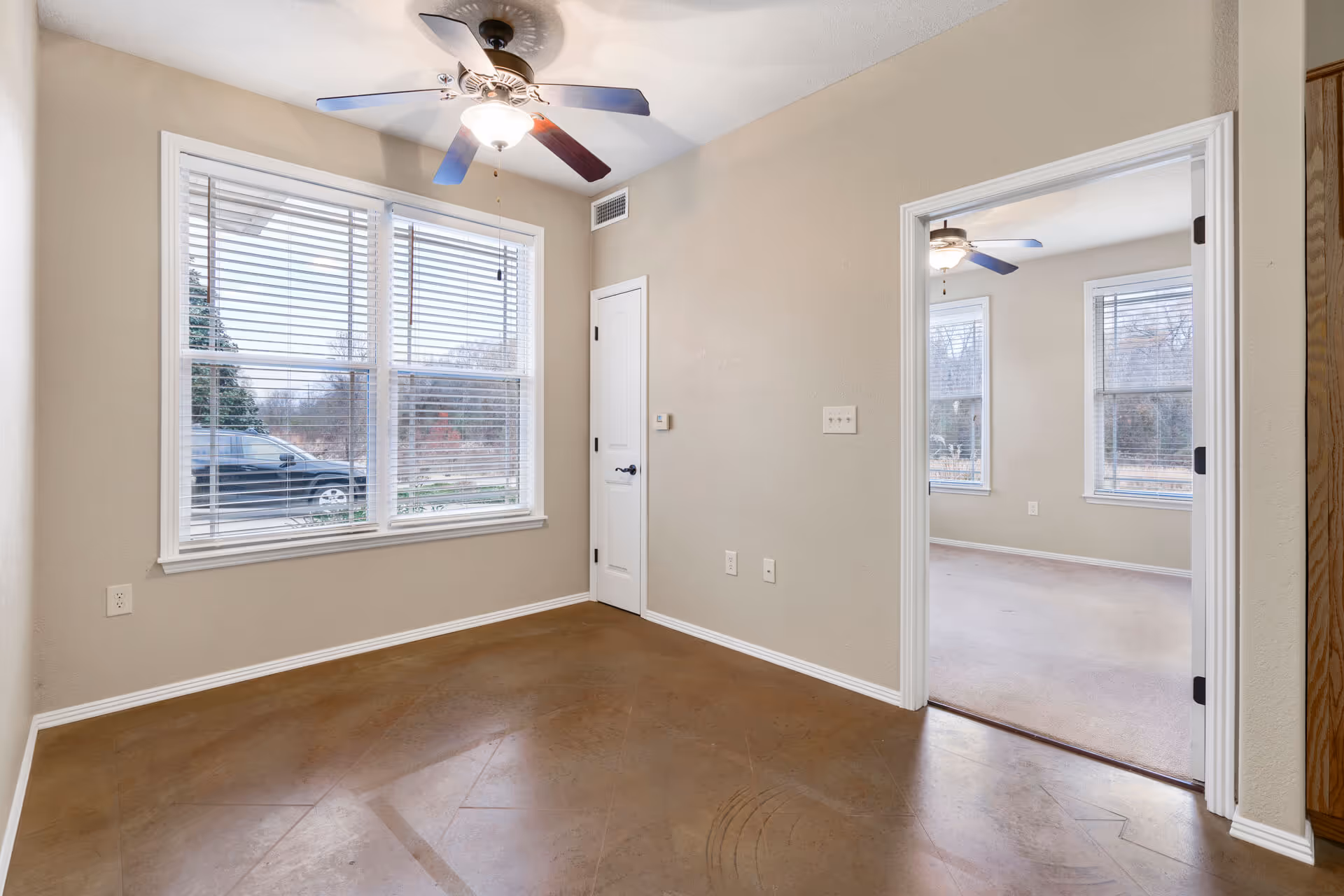 Empty interior room with a large window and blinds, a ceiling fan, and a doorway opening into an adjacent room.