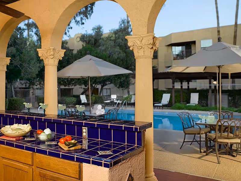 Outdoor patio area with a tiled counter and wooden cabinets under a covered structure with arches. On the counter are bowls with chips, tomatoes, and other snacks. In the background, there is a swimming pool surrounded by lounge chairs and tables with umbrellas, set in a garden-like environment with trees and a multi-story building.