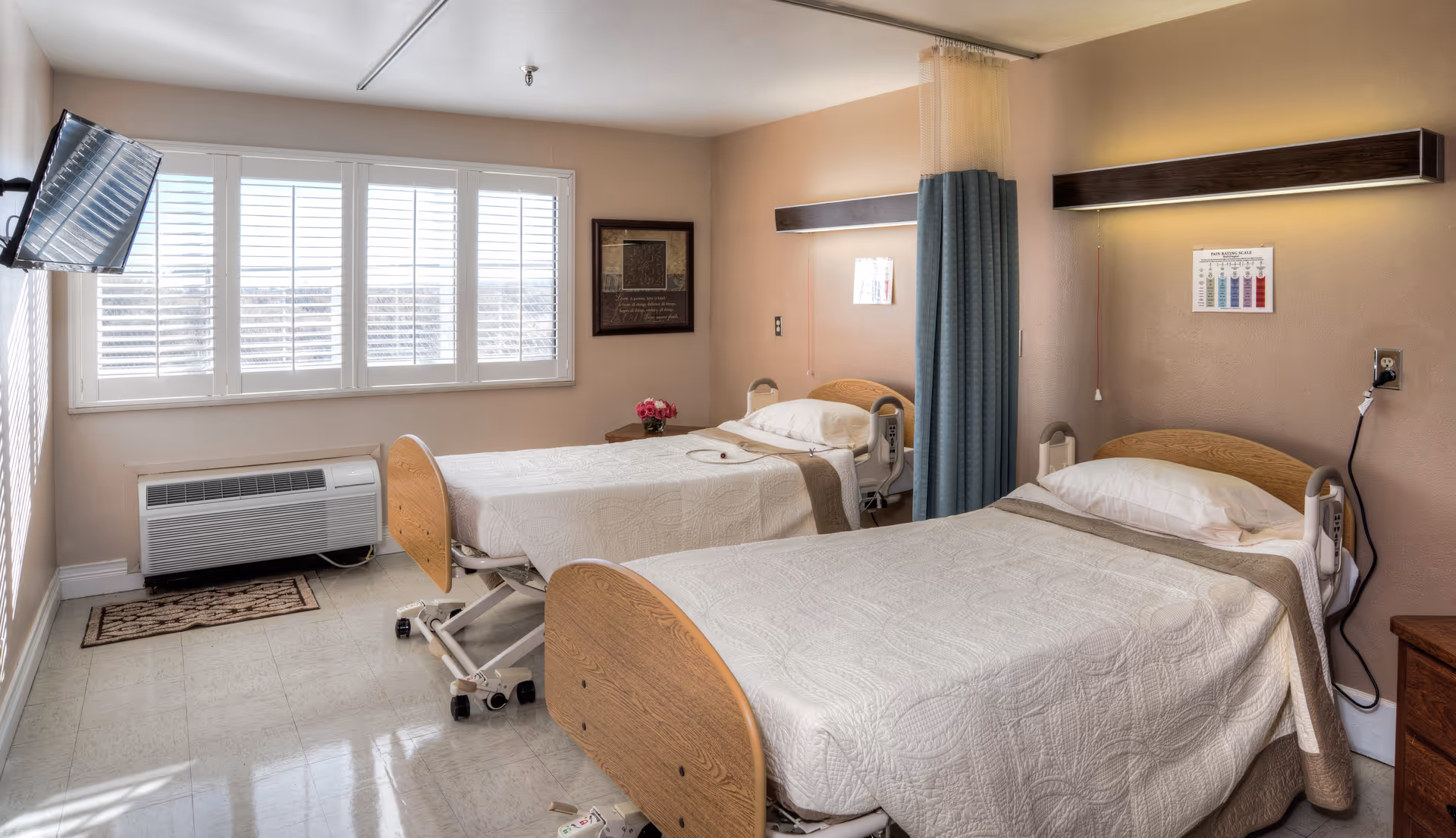 A healthcare facility room with two hospital beds, each made with white quilts and pillows. There is a blue privacy curtain between the beds, a wall-mounted TV on the left, a window with white shutters, and a framed picture on the wall. The room has beige walls and a tiled floor.