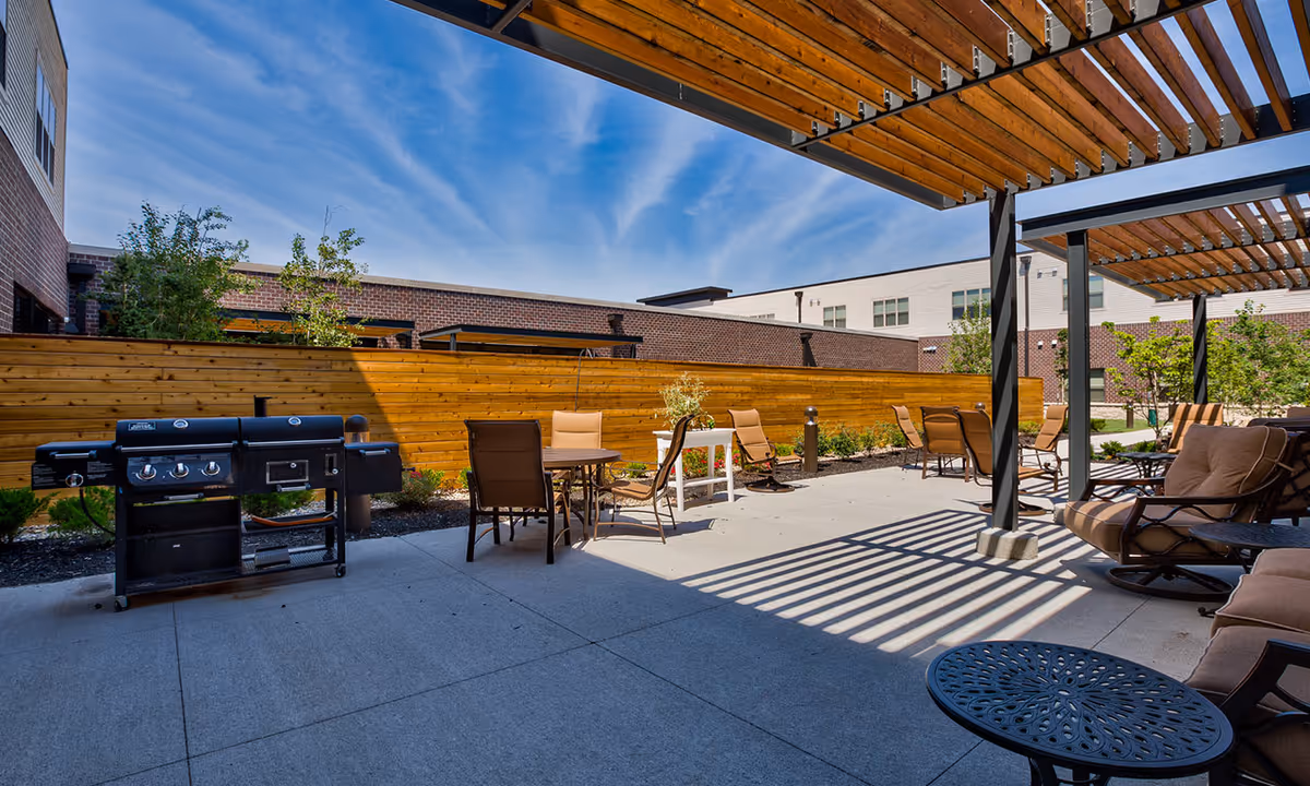 Outdoor patio area at Silvercrest at College View Senior Living featuring a large grill, several tables with chairs, cushioned lounge chairs under a pergola, and a wooden privacy fence with plants and trees in the background under a blue sky.