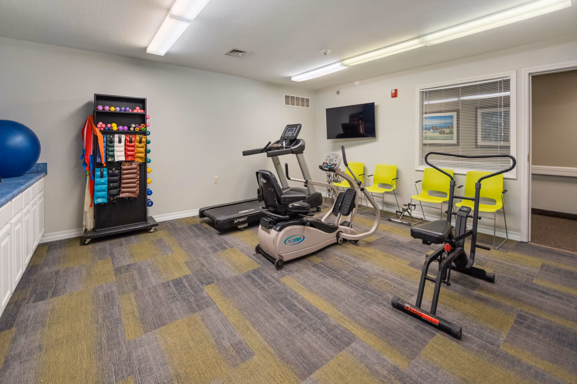 A small fitness room with exercise equipment including a recumbent bike, treadmill, and a seated exercise machine. There is a rack with colorful exercise bands and balls on the left side, a blue exercise ball on a countertop, and four yellow chairs lined up against the wall beneath a mounted flat-screen TV. The room has gray and green patterned carpet and white walls.