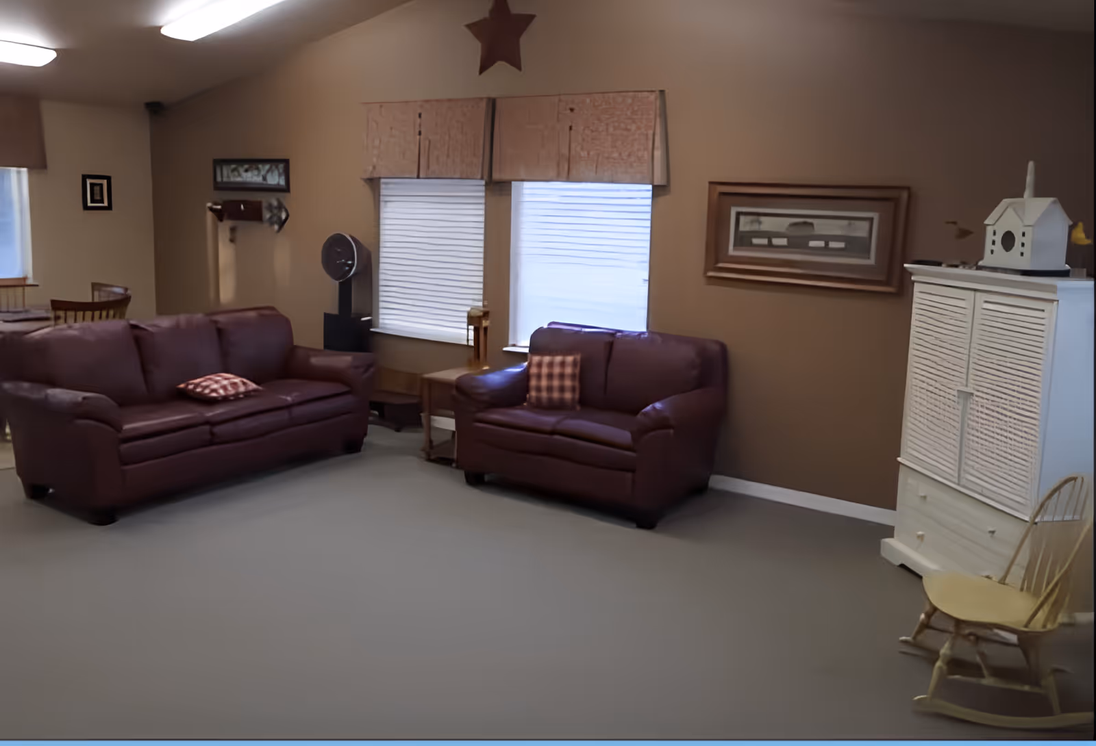 A cozy living room with two maroon leather sofas, each with a patterned cushion. There are two windows with blinds and valances on the wall behind the sofas. A small wooden side table is placed between the sofas. On the right side, there is a white cabinet with a decorative birdhouse on top and a wooden rocking chair next to it. The walls are painted beige and decorated with framed artwork and a star above the windows.