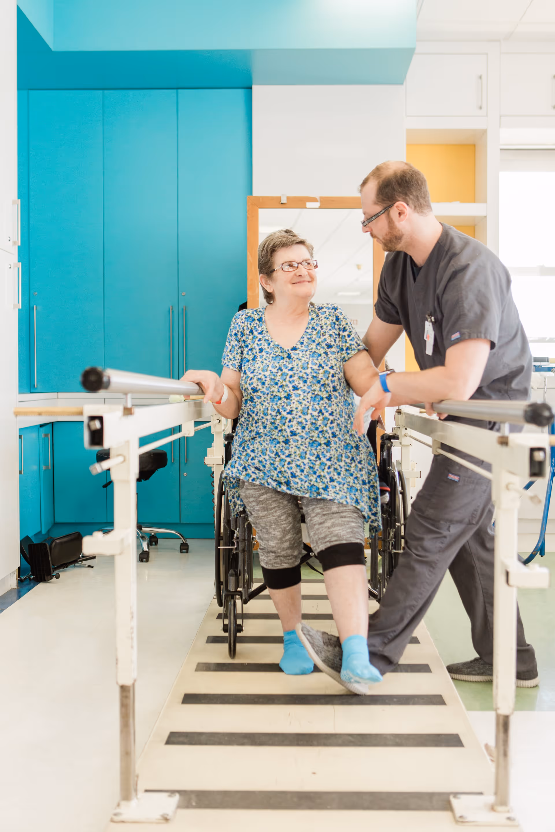 A healthcare professional assists an elderly woman with physical therapy as she practices walking with support bars in a rehabilitation facility. The woman is stepping forward with one leg while holding onto the bars, and the professional is providing support and encouragement.