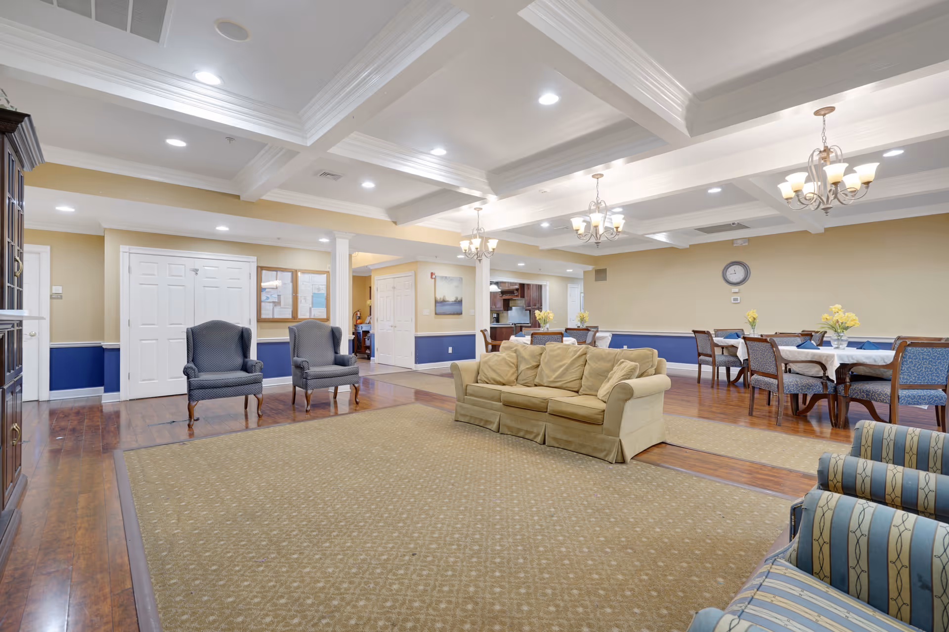 A spacious common area in a senior living facility featuring a beige couch, two blue armchairs, and a patterned carpet. In the background, there are several dining tables with chairs, each table decorated with yellow flower arrangements. The room has a coffered ceiling with recessed lighting and chandeliers, wooden flooring, and walls painted in beige and blue tones.