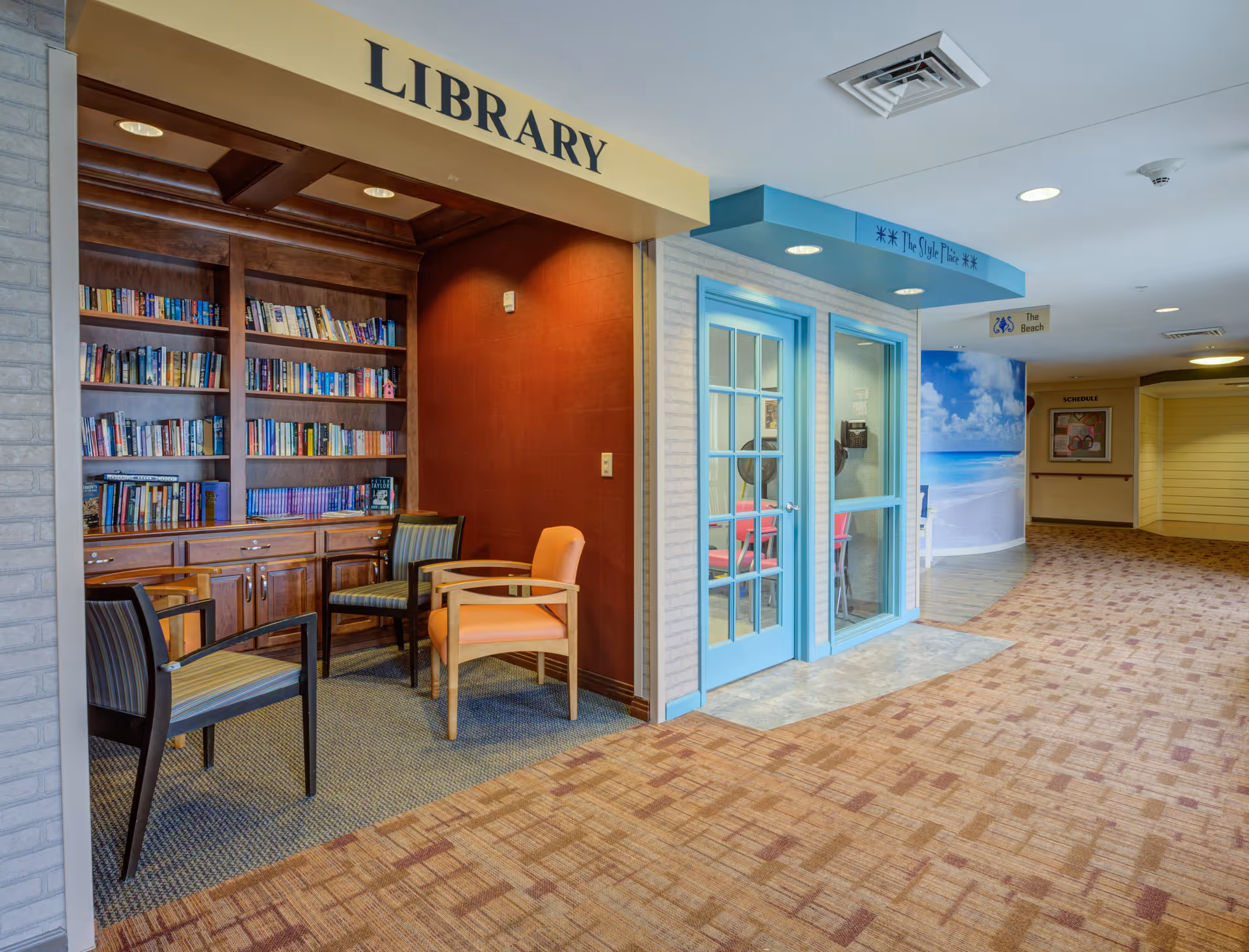 Interior view of a senior living facility hallway featuring a small library nook with wooden bookshelves filled with books and three chairs. Adjacent to the library is a room with blue-framed glass doors labeled 'The Style Place.' The hallway has carpeted flooring and signs indicating directions to 'The Beach' and a schedule board further down the corridor.
