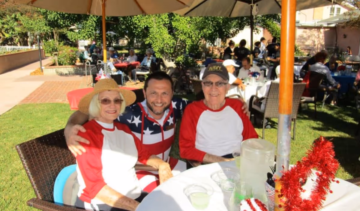 Three people sitting at a round outdoor table under an umbrella in a garden setting. Two elderly individuals wearing matching red and white shirts are seated on either side of a younger man dressed in a star-spangled outfit. Other people are visible in the background, also seated at tables, enjoying the sunny day.