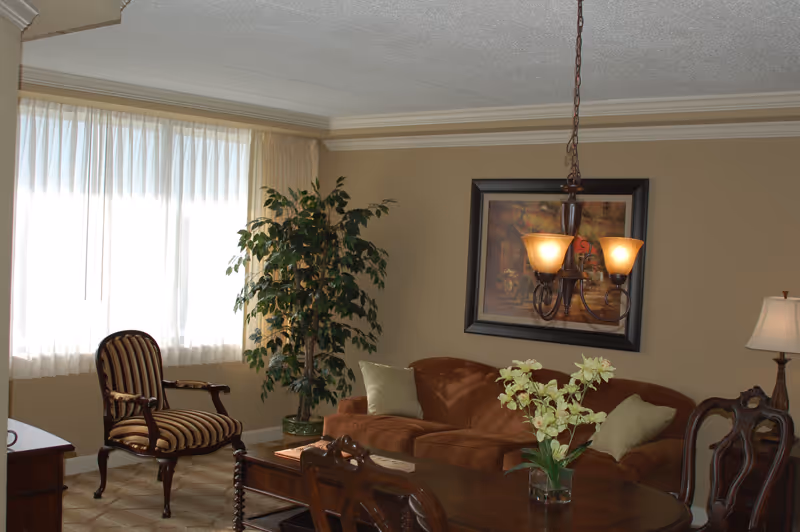 A cozy living room with a brown sofa adorned with light-colored pillows, a wooden coffee table with a vase of flowers, a striped armchair near a window with sheer curtains, a tall green plant in the corner, a framed painting on the beige wall, and a hanging light fixture with three lamps.