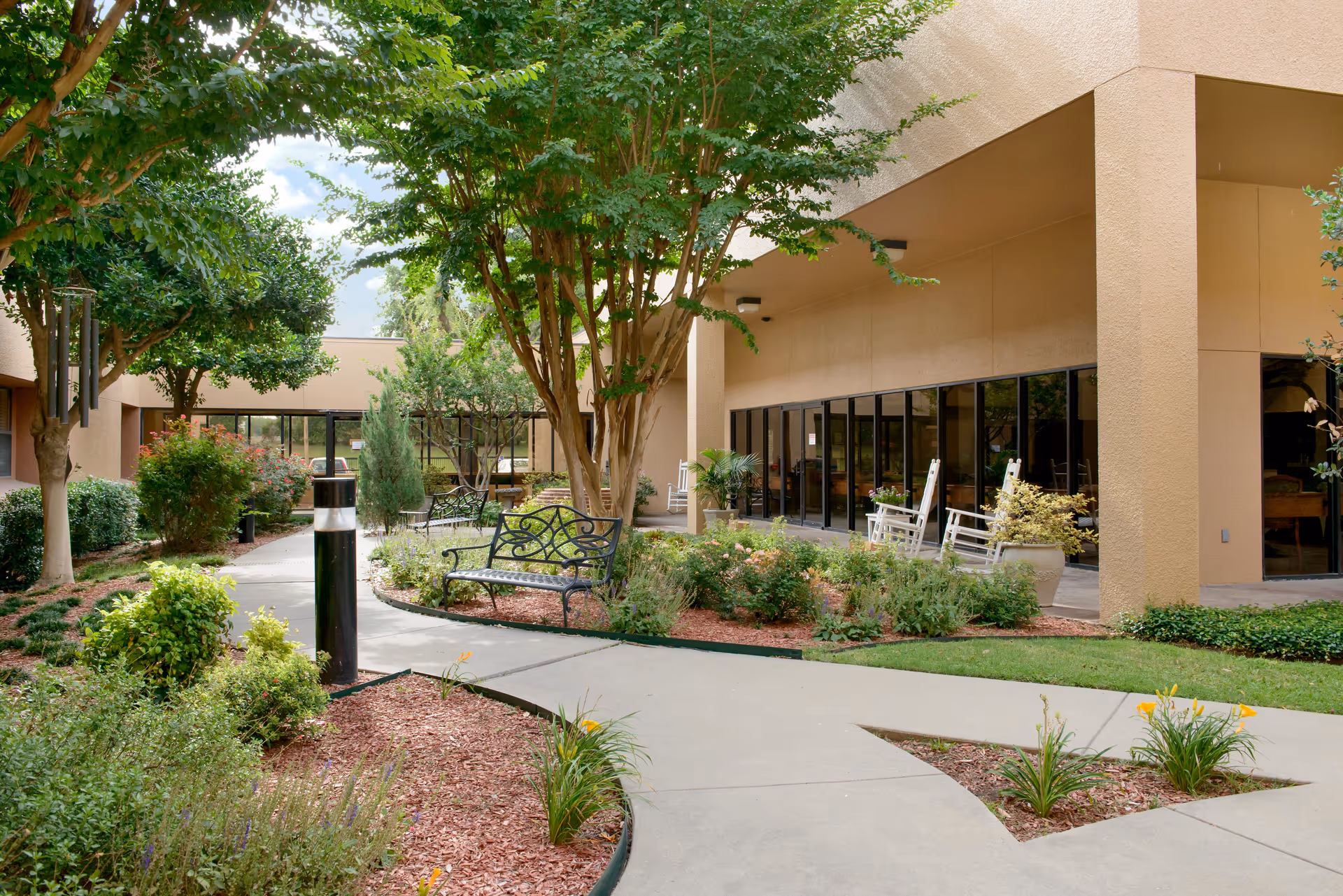 Outdoor courtyard area at Renaissance Park Multi Care Center featuring a paved walkway, green trees, bushes, flower beds with mulch, black metal benches, and white rocking chairs on a covered patio next to the building.