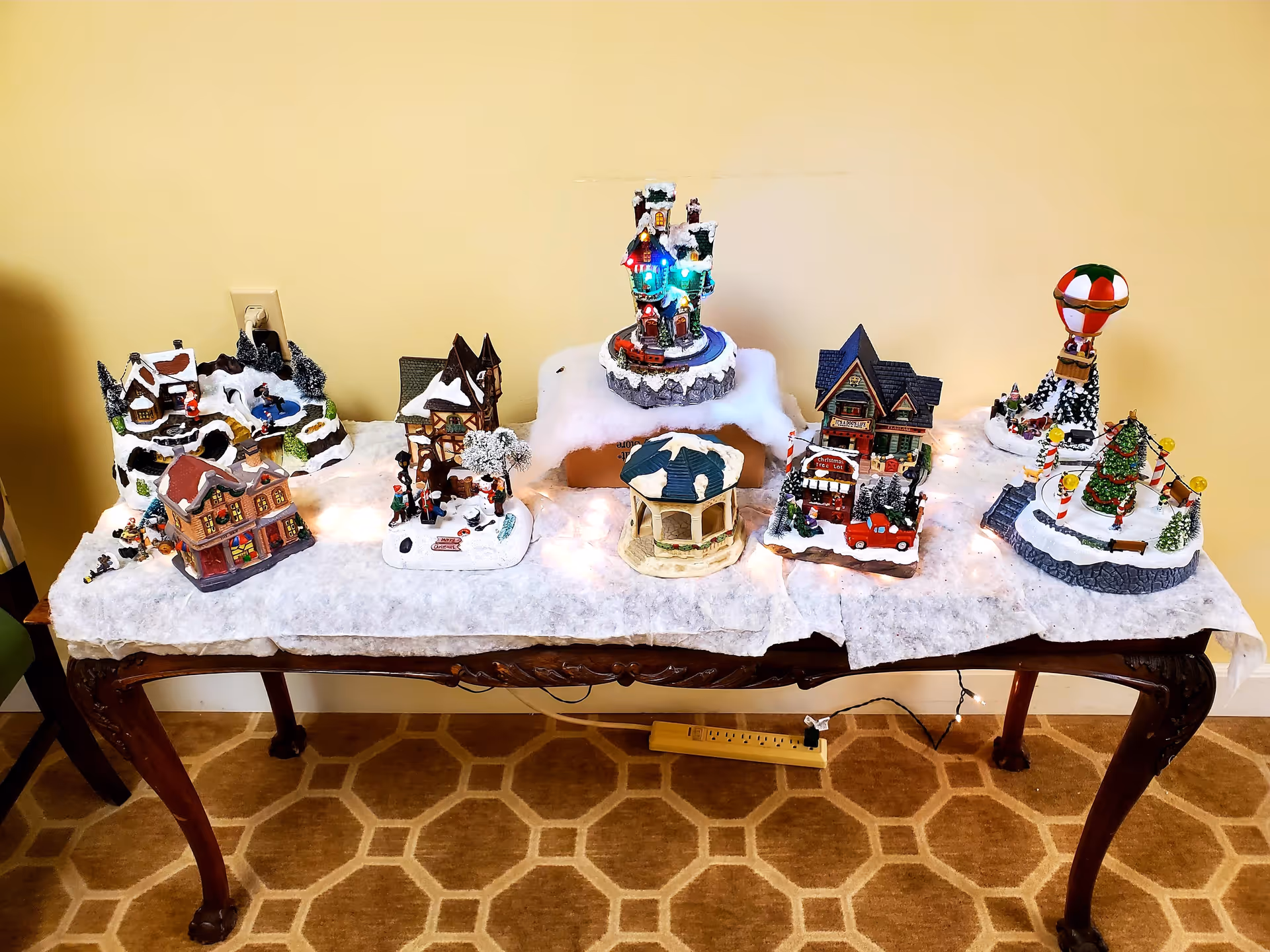 A decorative Christmas village display on a wooden table covered with white fabric resembling snow. The display includes miniature houses, a gazebo, a hot air balloon, and a Christmas tree with string lights. The background is a plain beige wall and the floor has a patterned carpet.
