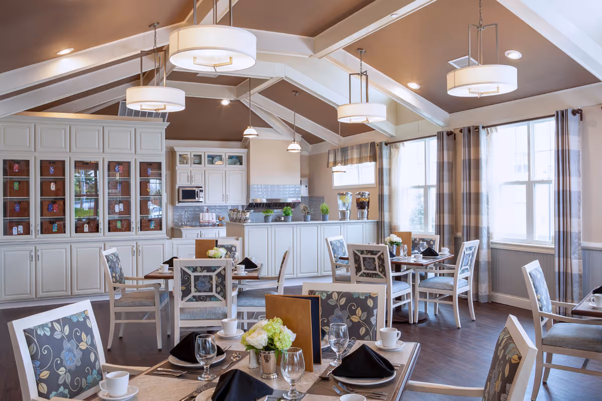 A bright and elegant dining room with several tables set with plates, glasses, cups, and black napkins. The room features floral-patterned chairs, large windows with plaid curtains, white cabinetry, and a counter with beverage dispensers. The ceiling has exposed beams and modern hanging light fixtures.
