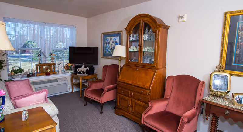 A cozy living room with two pink upholstered armchairs, a wooden cabinet with glass doors displaying decorative items, a small TV on a wooden side table, a window with lace curtains, and a floral-patterned sofa with pink cushions.