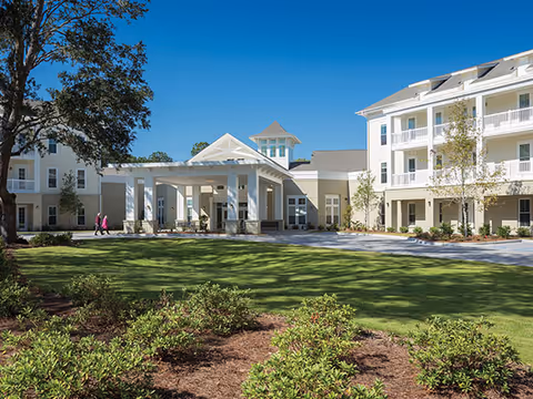 Front exterior of a three-story senior living building with a covered entrance, balconies, landscaped lawn, and two people near the entry under a clear blue sky.