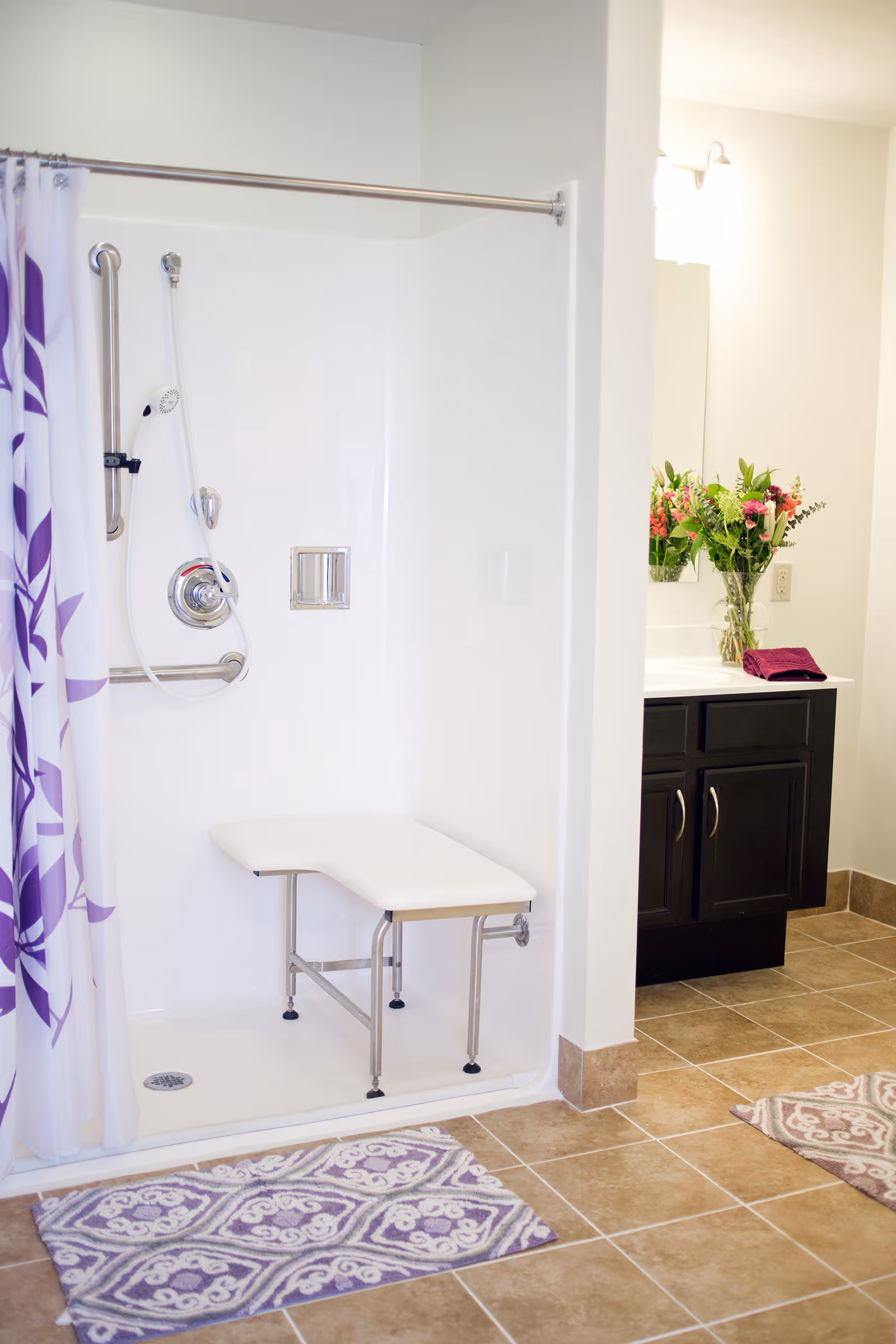 A clean bathroom featuring a walk-in shower with a white fold-down bench and a handheld showerhead. The shower has a purple and white patterned curtain. Adjacent to the shower is a vanity with a dark wood cabinet, a white countertop, a mirror, a vase of colorful flowers, and a folded purple towel. The floor is tiled with brown tiles and has two purple and white patterned rugs.