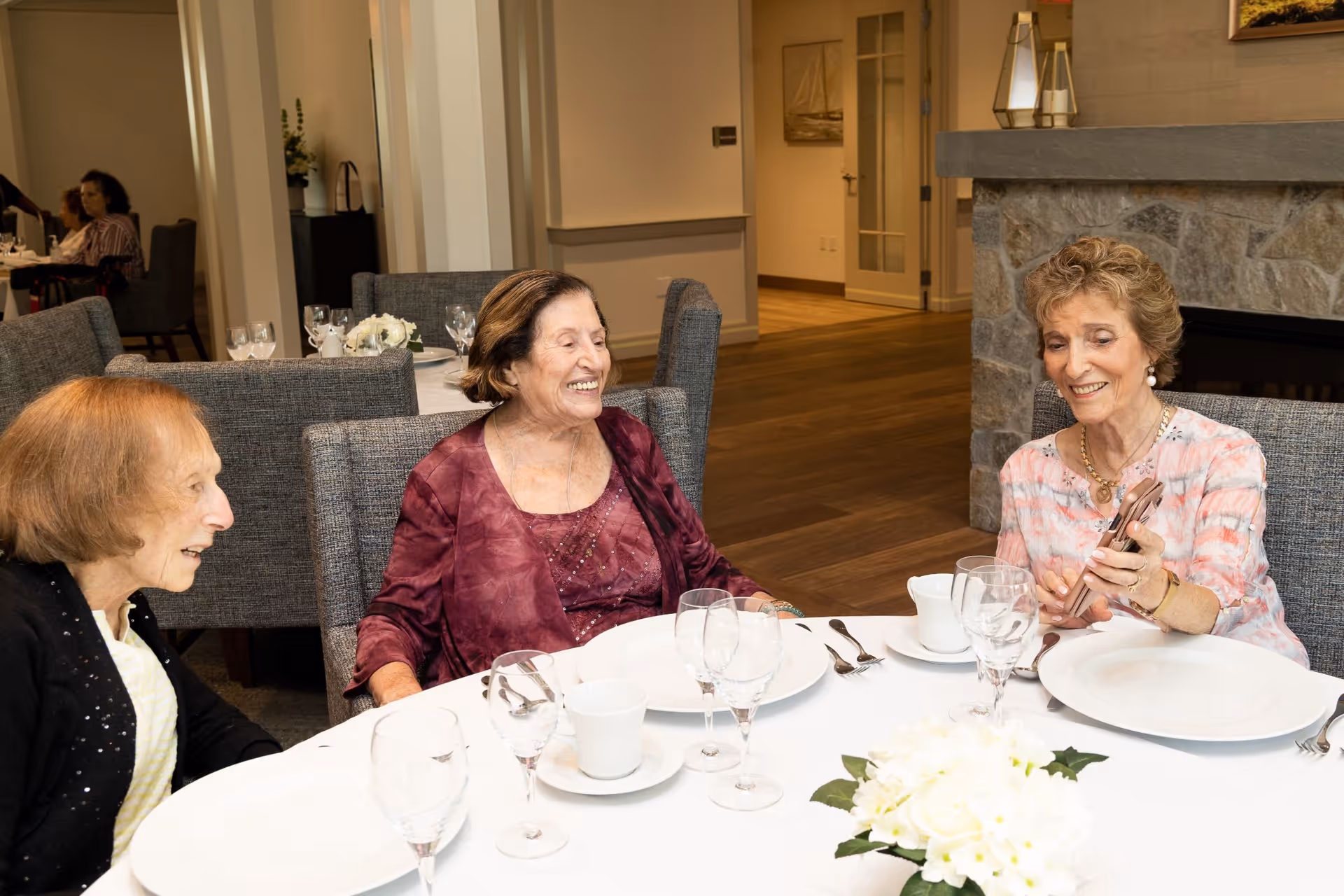 Three elderly women sitting around a dining table in a well-lit room, smiling and engaging with each other. The table is set with white plates, cups, and glasses, and there is a white floral centerpiece. The background shows more dining chairs and a stone fireplace.