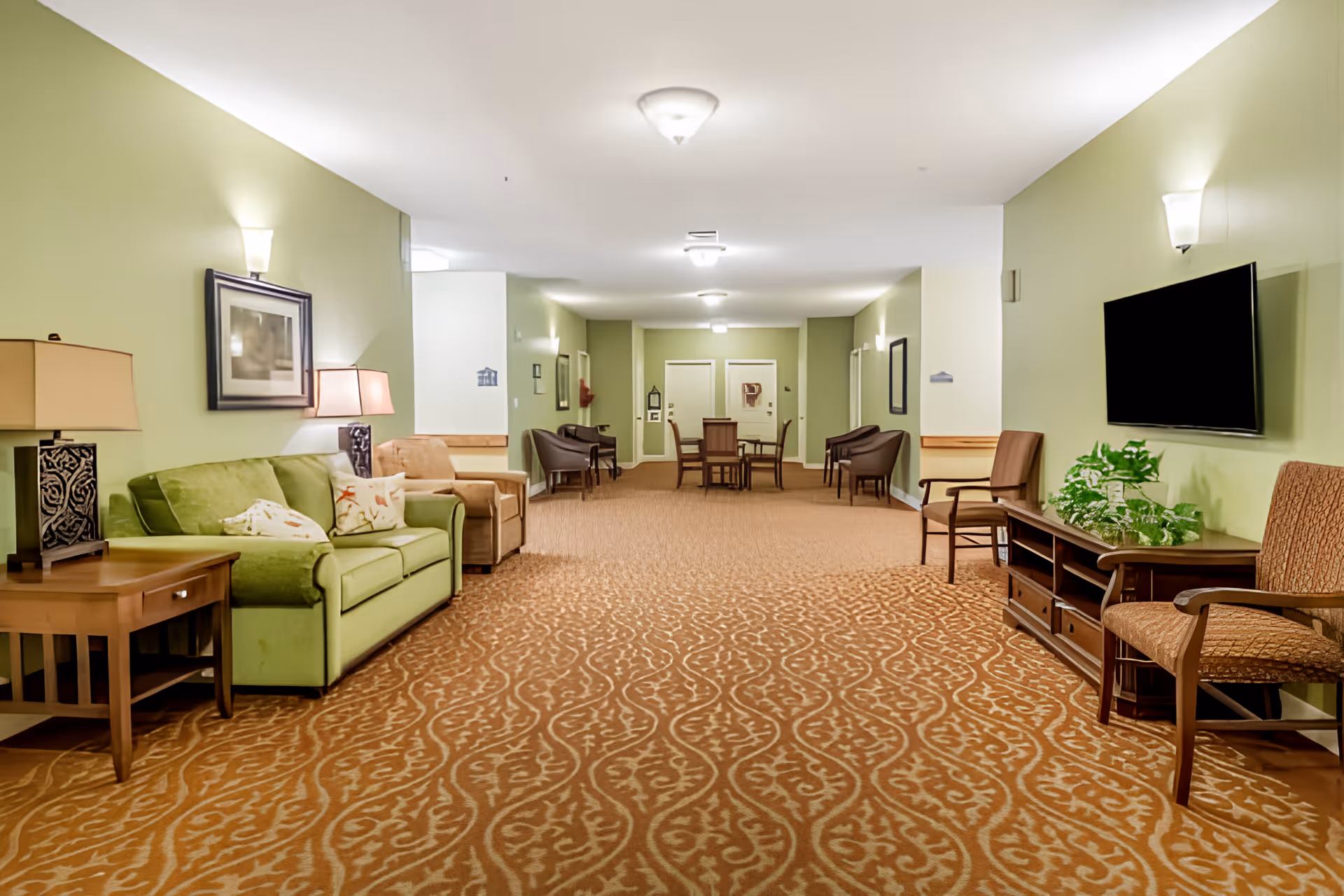 Spacious, well-lit common lounge area with sofas, chairs, tables, a wall-mounted TV and patterned carpet in an assisted living facility.